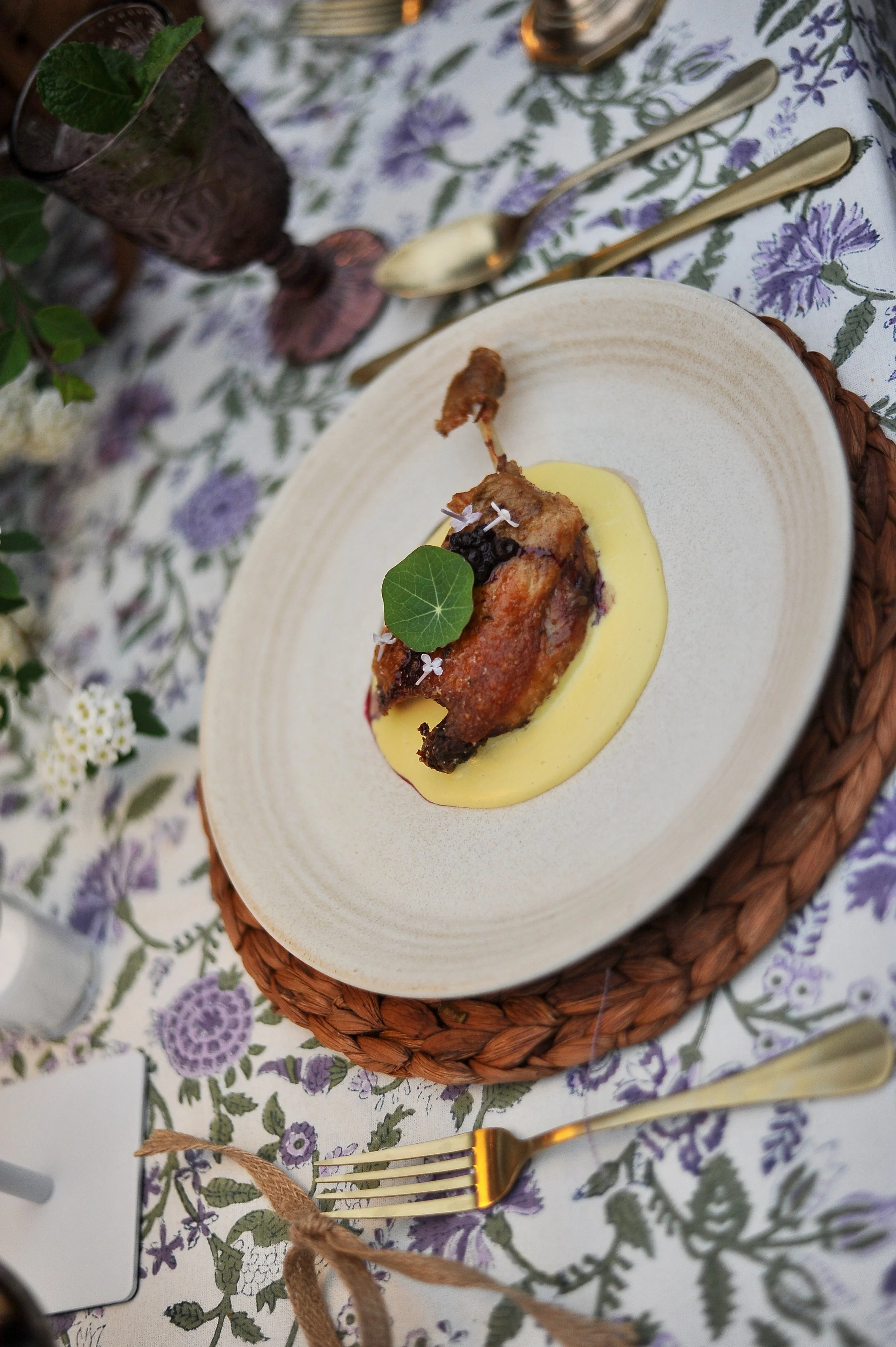 A plated roasted chicken leg on a bed of creamy yellow sauce, garnished with a green leaf and small white flowers, set on a decorative tablecloth with purple and green floral pattern, surrounded by gold-colored cutlery and a purple vase with green fo