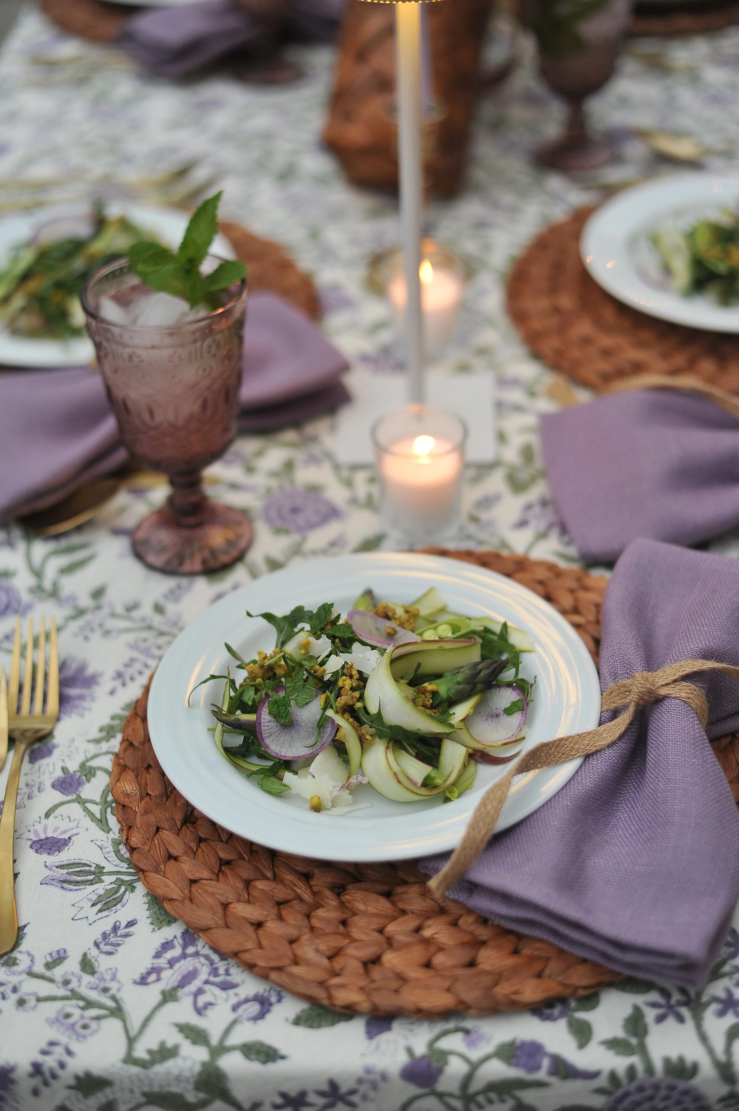A dinner table setting with a white plate of mixed greens salad garnished with radish slices and asparagus, purple napkins tied with twine, purple glasses, candles, and a floral tablecloth with lavender and green patterns.