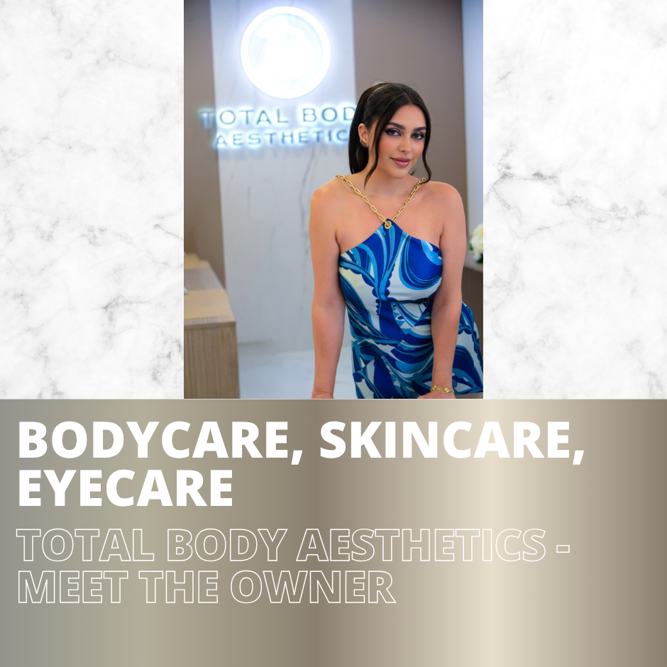 A woman wearing a blue patterned dress and gold chain necklace posing indoors in front of a sign that reads 'Total Body Aesthetics.'