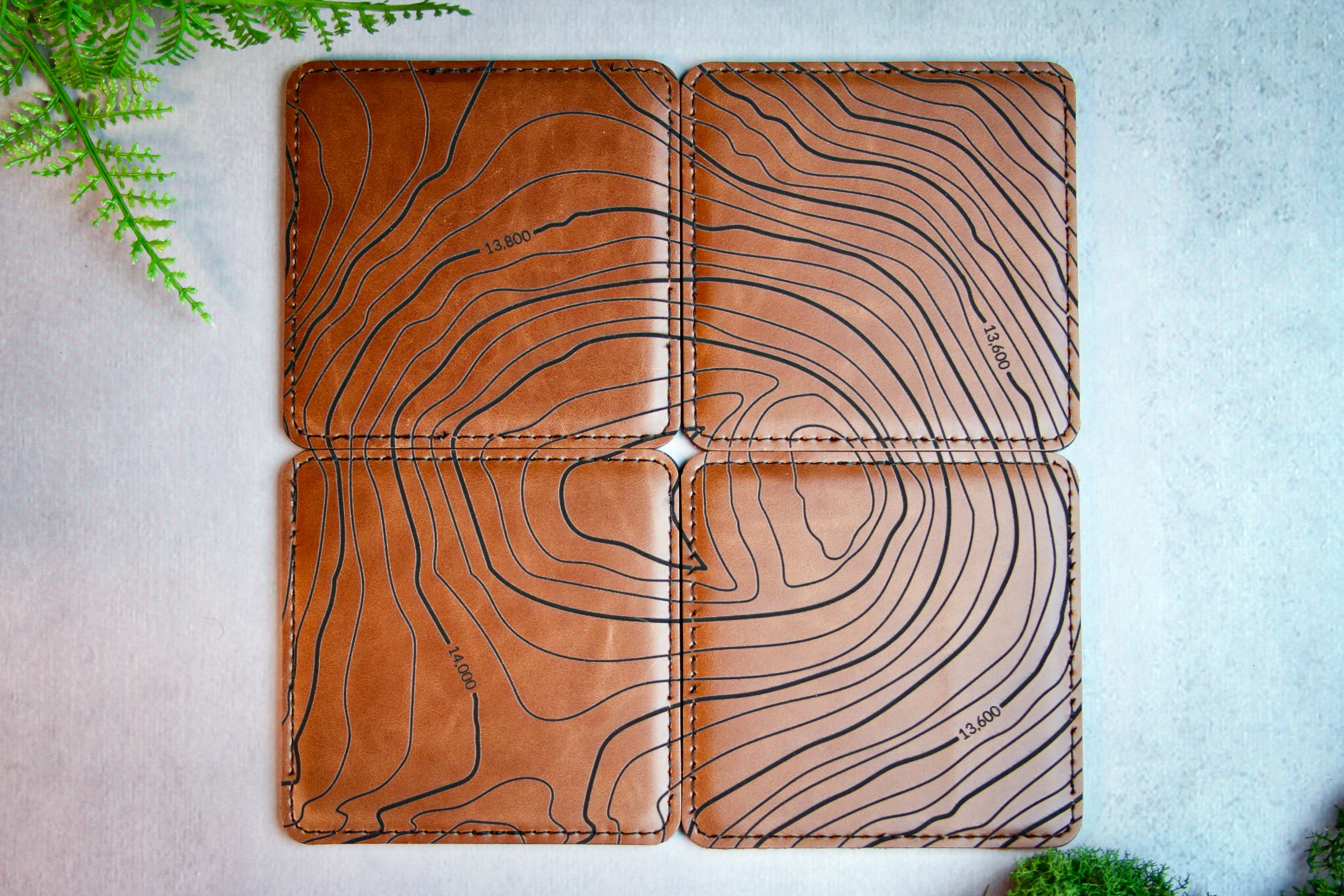 Square brown vegan leather coasters with topographic design labeled "Mount Rainier," displayed on a small wooden stand, with a coffee mug and jar of coffee beans in the background.