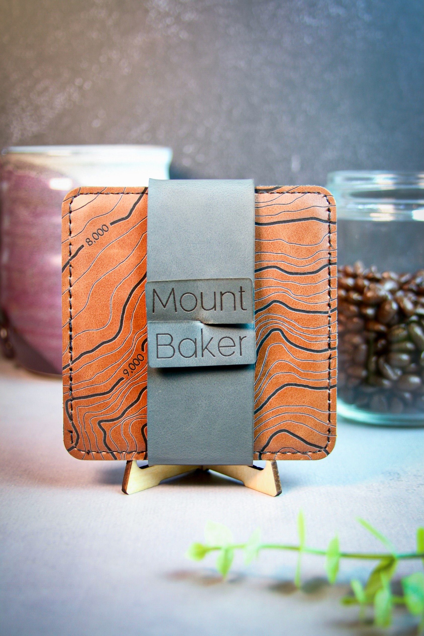 Square brown vegan leather coasters with topographic design labeled "Mount Baker," displayed on a small wooden stand, with a coffee mug and jar of coffee beans in the background.