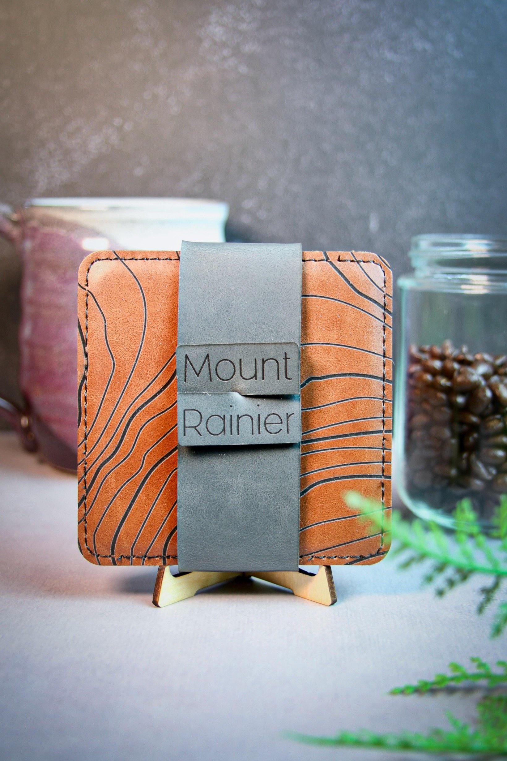 Square brown vegan leather coasters with topographic design labeled "Mount Rainier," displayed on a small wooden stand, with a coffee mug and jar of coffee beans in the background.
