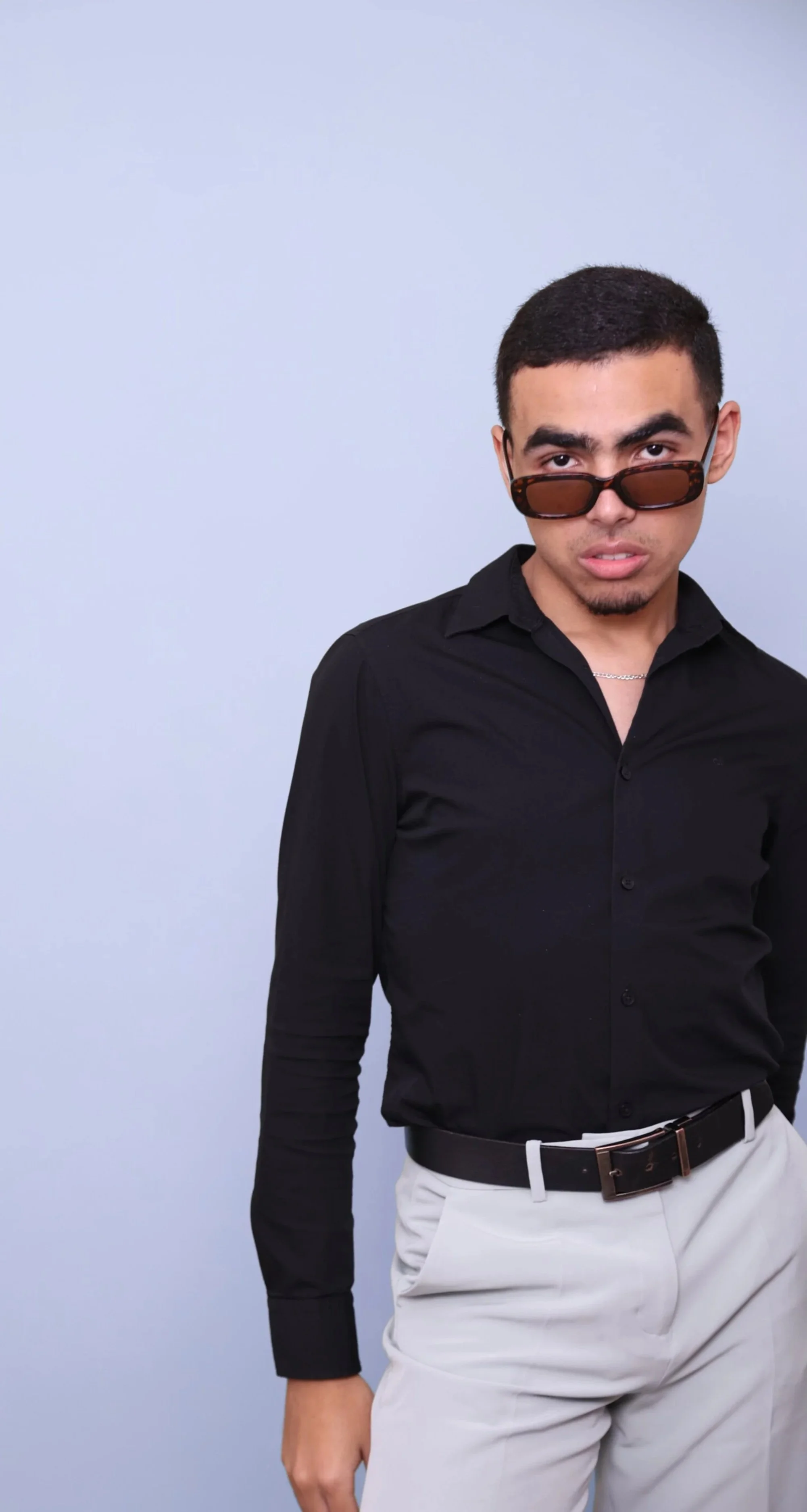 A young man wearing a black button-up shirt, white pants, and sunglasses, standing against a light gray background.