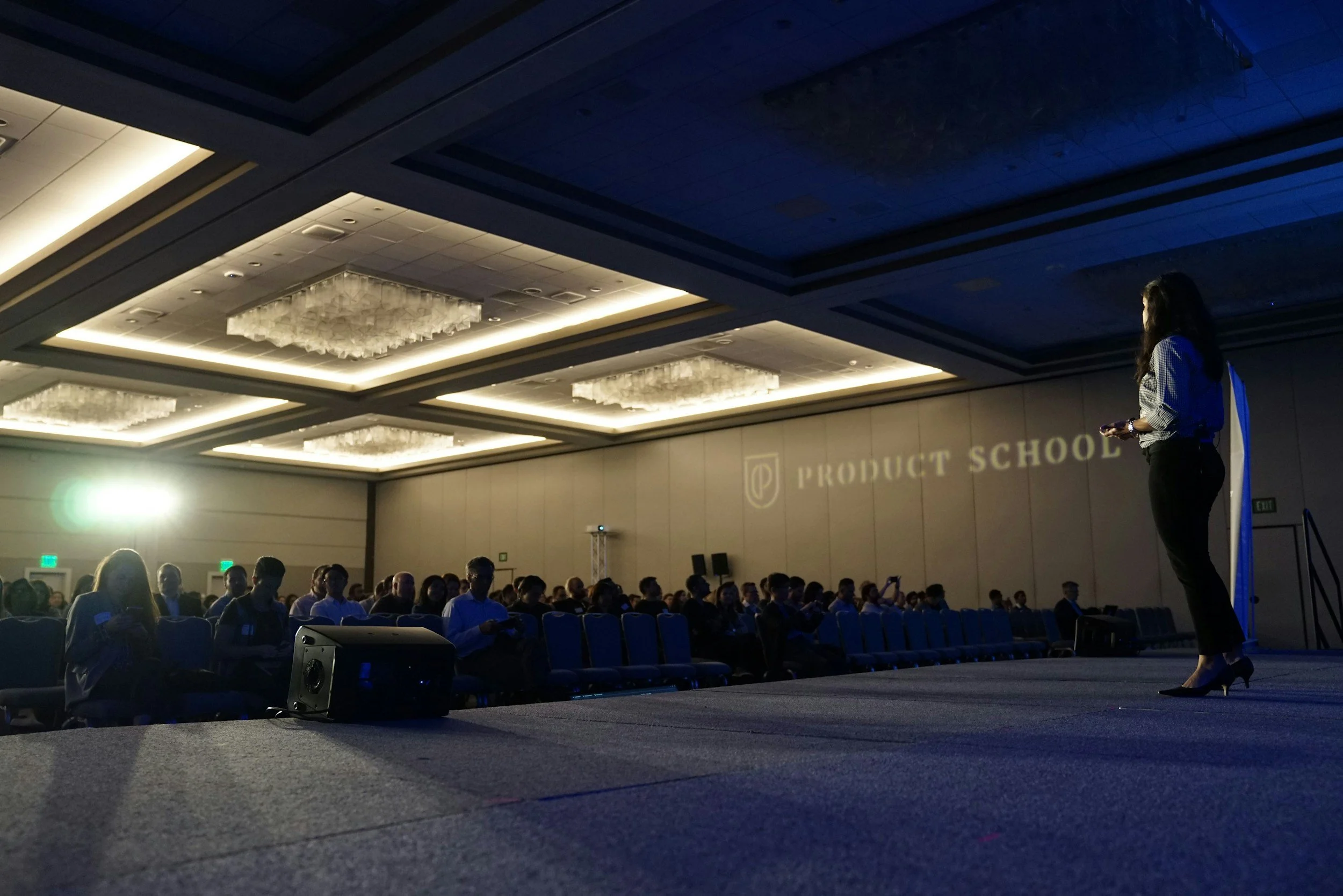 A woman is standing on a stage giving a presentation to an audience in a conference room. The room has a high ceiling with large chandeliers and the projection on the wall says 'Product School'.