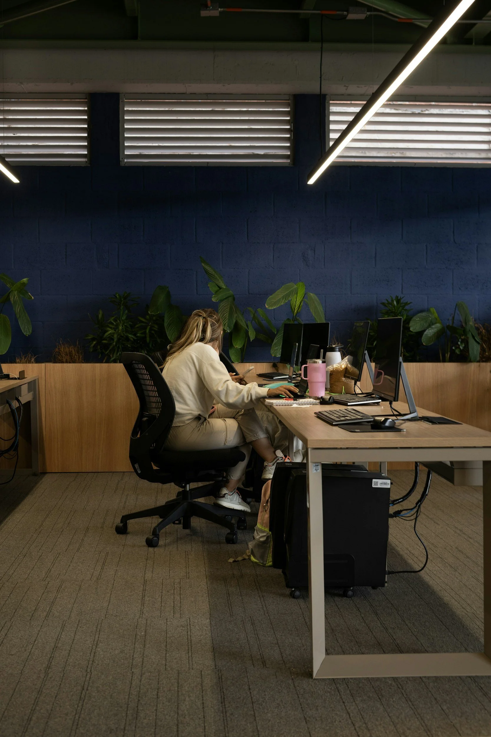 An office worker sitting at a desk with multiple computer monitors, plants in the background, and a dark blue wall behind.