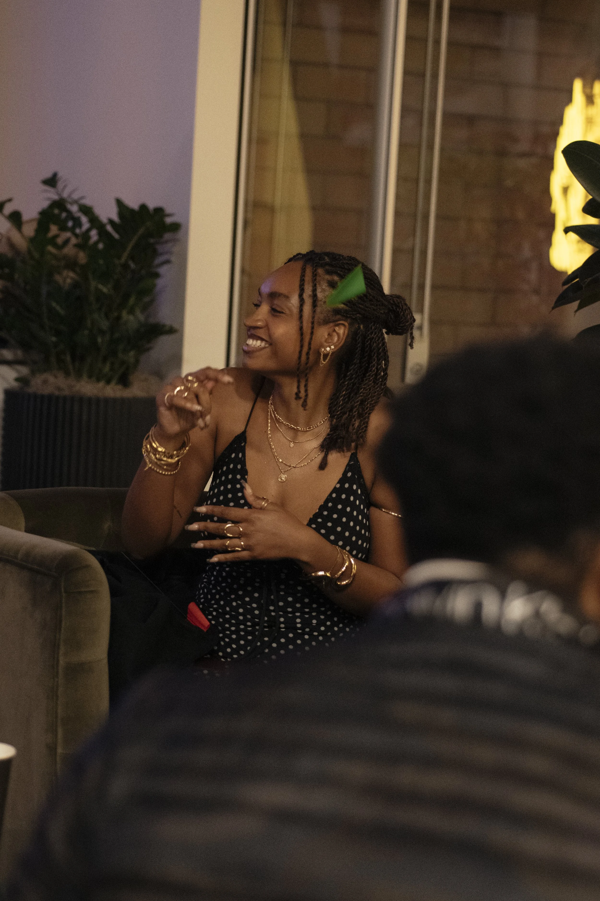 A woman with curly hair in braids, wearing a black polka dot dress and gold jewelry, laughing and enjoying herself at a social gathering.