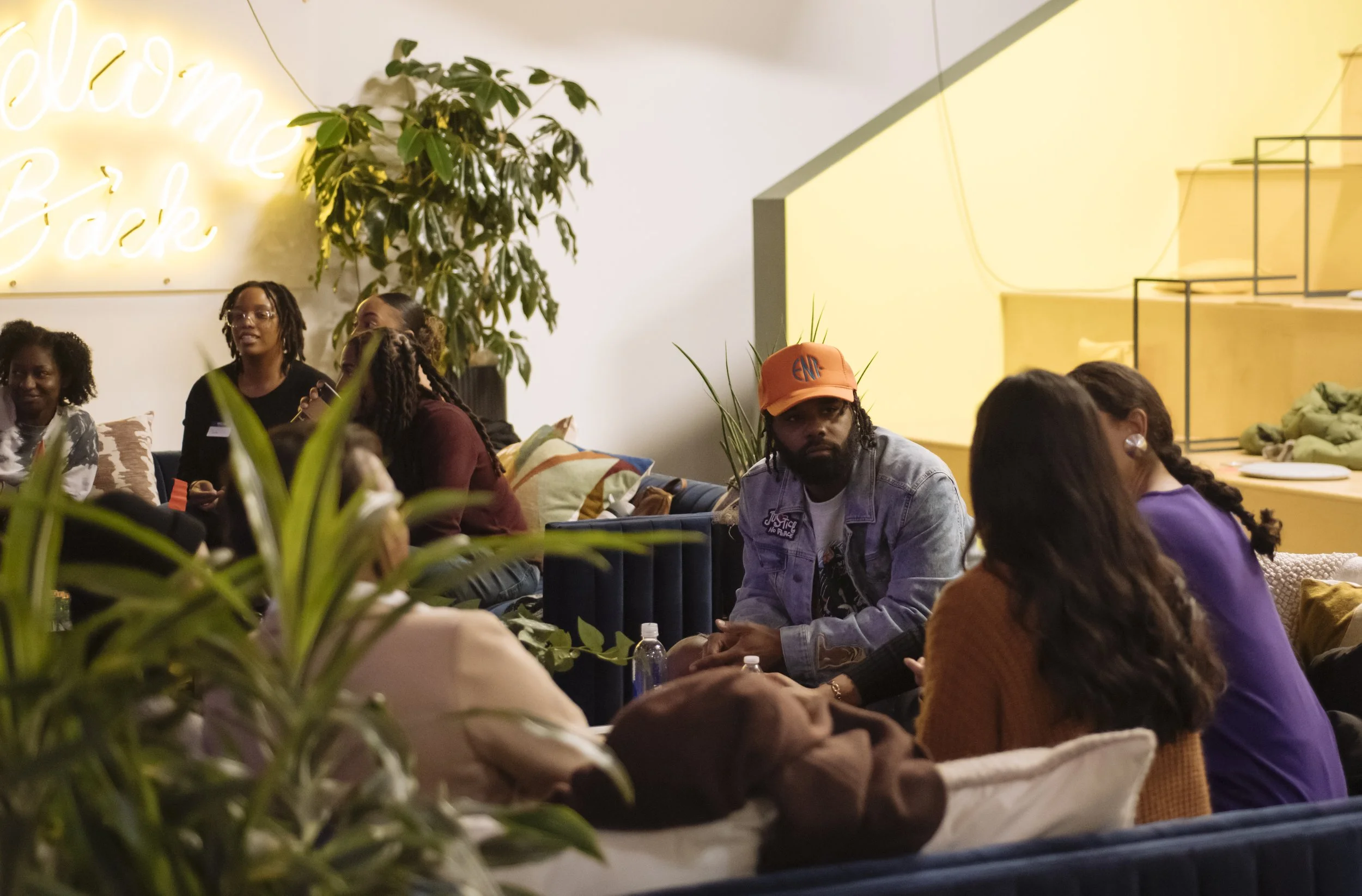 Group of people sitting and talking in a cozy, modern indoor space with plants and a neon sign that reads 'Welcome Back' in the background.
