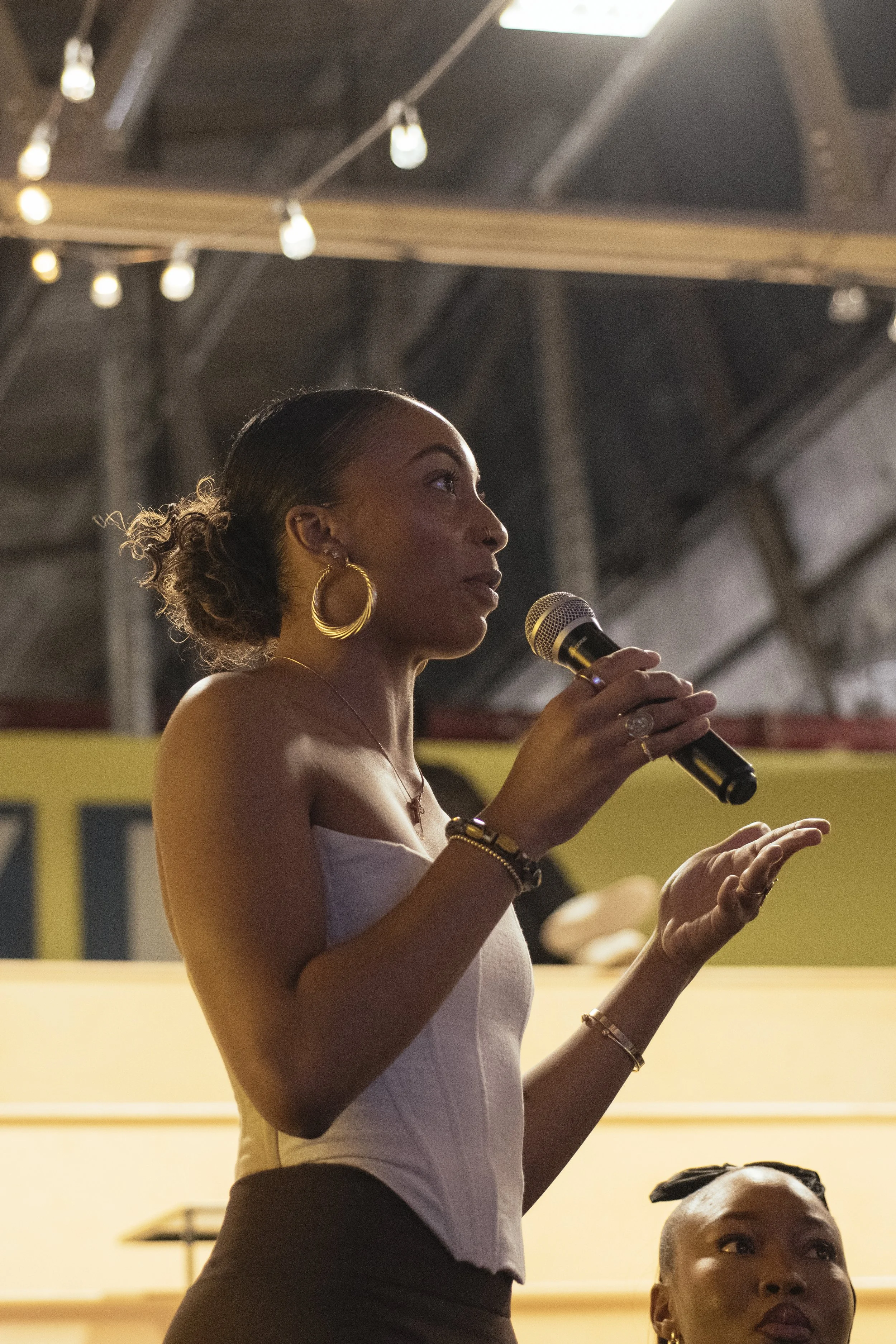 A woman is speaking into a microphone, wearing a strapless top, large gold hoop earrings, and various bracelets. She has her hair styled in a bun with curly ends, and appears to be in an indoor event space with string lights hanging overhead.