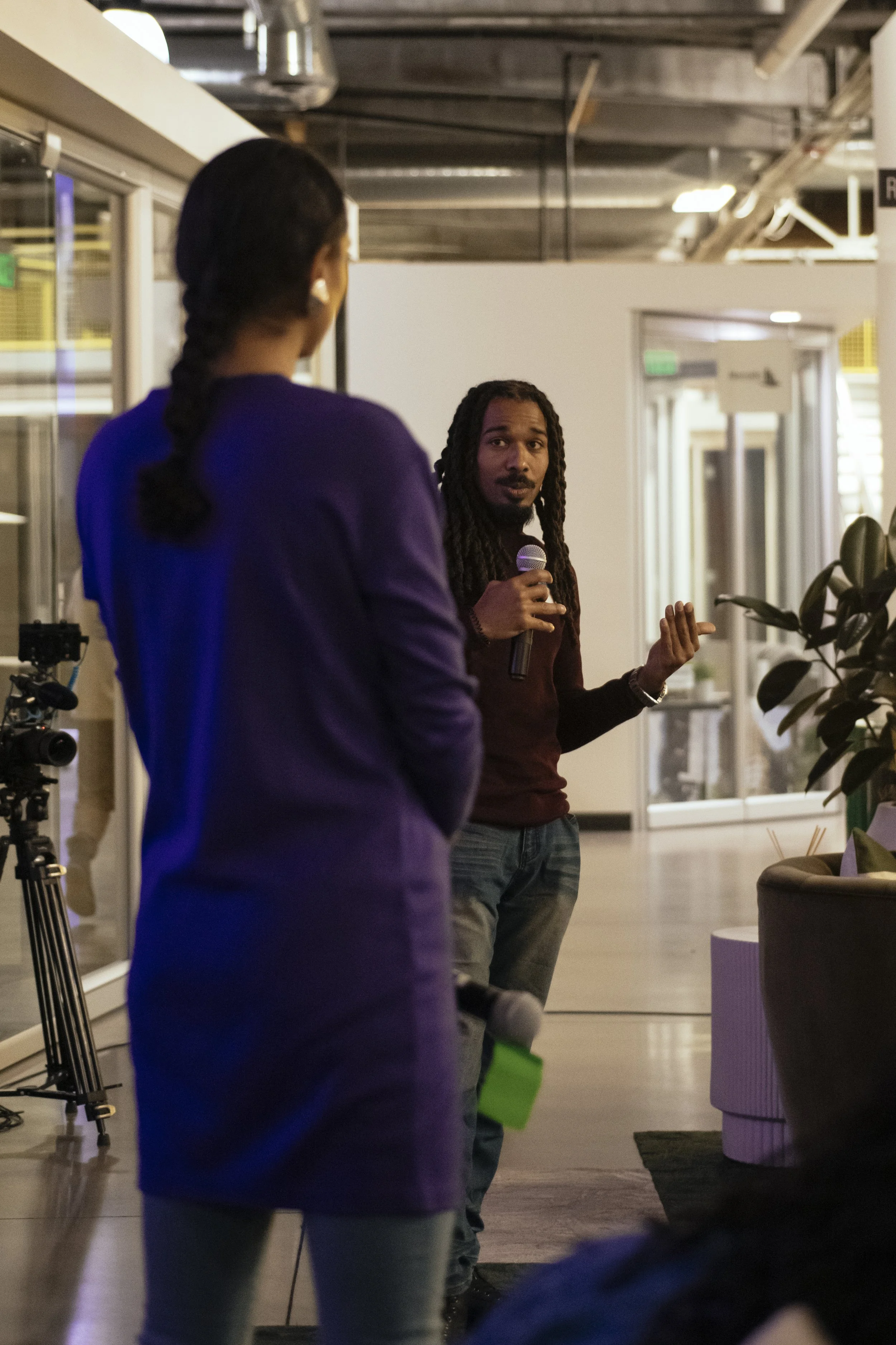 A man with long dreadlocks holding a microphone and speaking to a woman with a braid, in a modern indoor setting with glass doors and a camera on a tripod nearby.