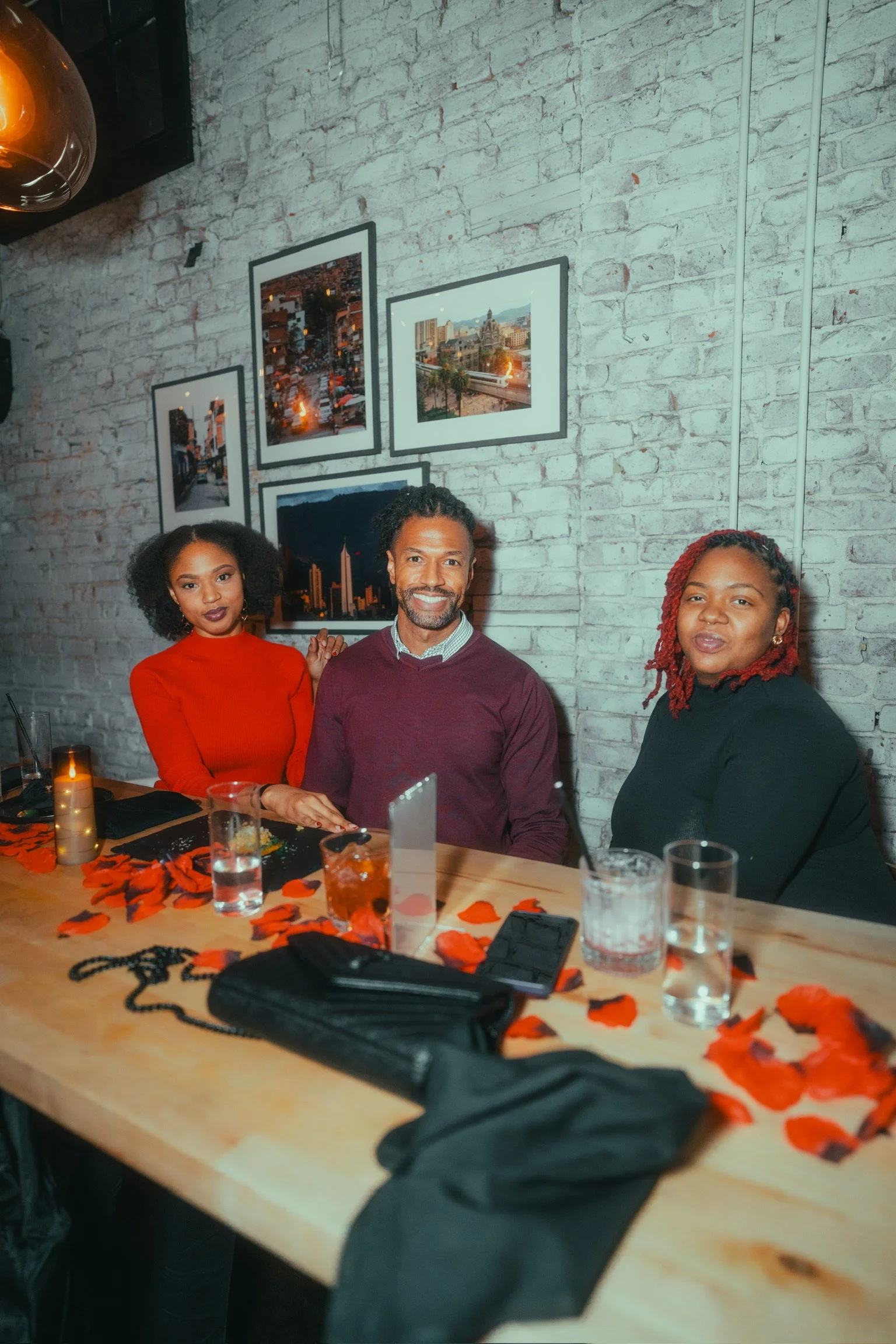 Three people sitting at a table decorated with red rose petals in a restaurant or cafe with brick walls and framed cityscape photos.