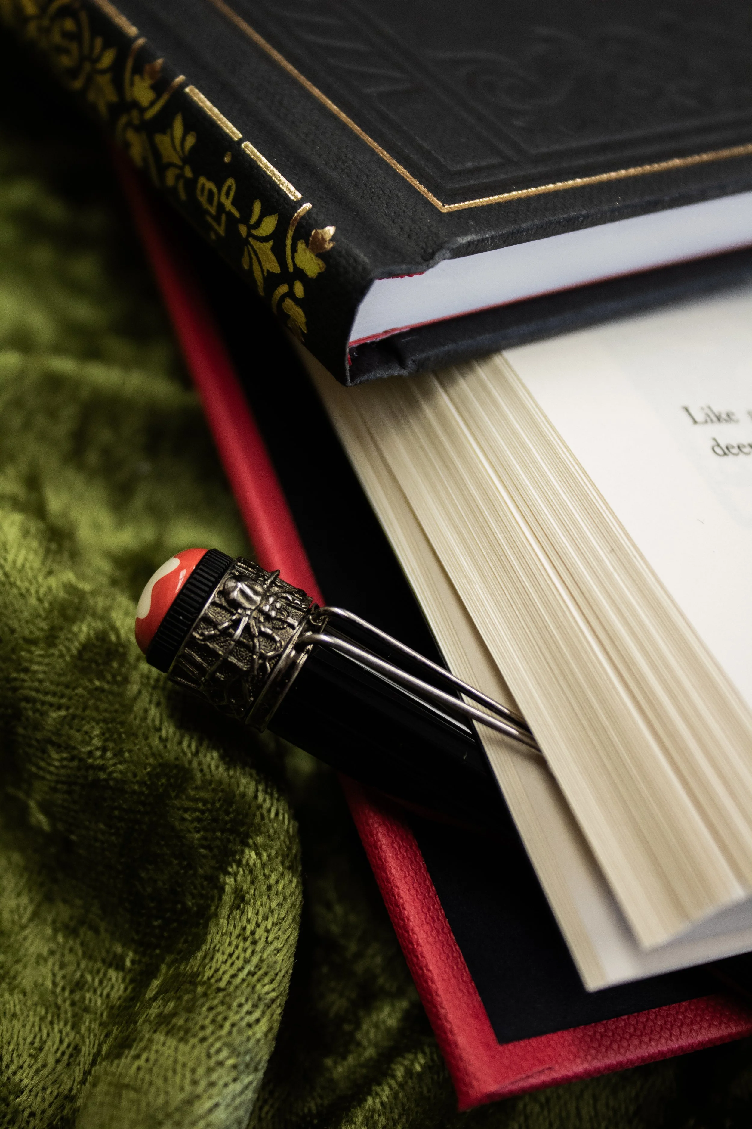 Close-up of a black and red fountain pen placed between two stacked hardcover books with ornate covers, resting on a green textured fabric.