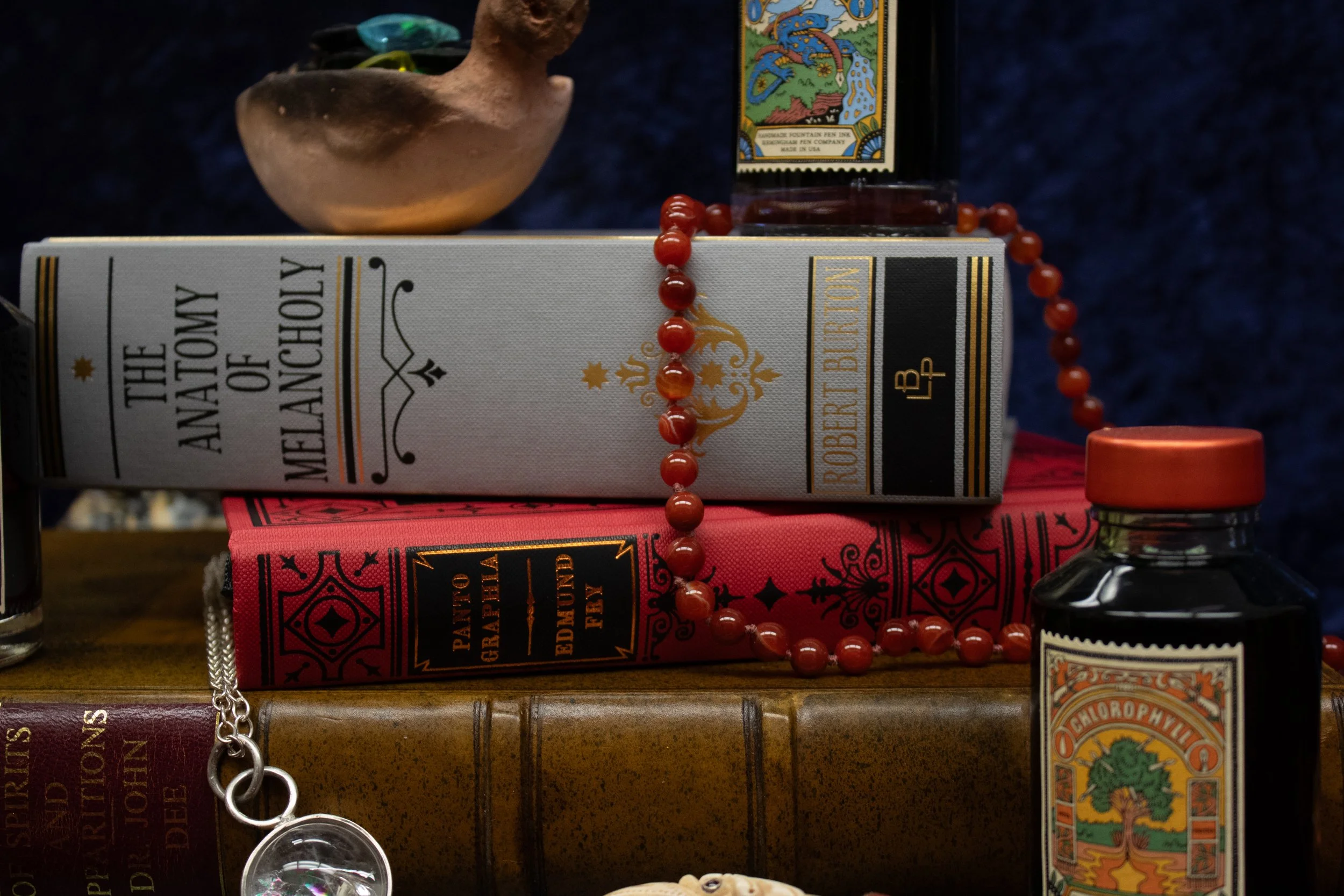 Stack of vintage books with a decorative rosary and small vials, featuring "The Anatomy of Melancholy" by Robert Burton.