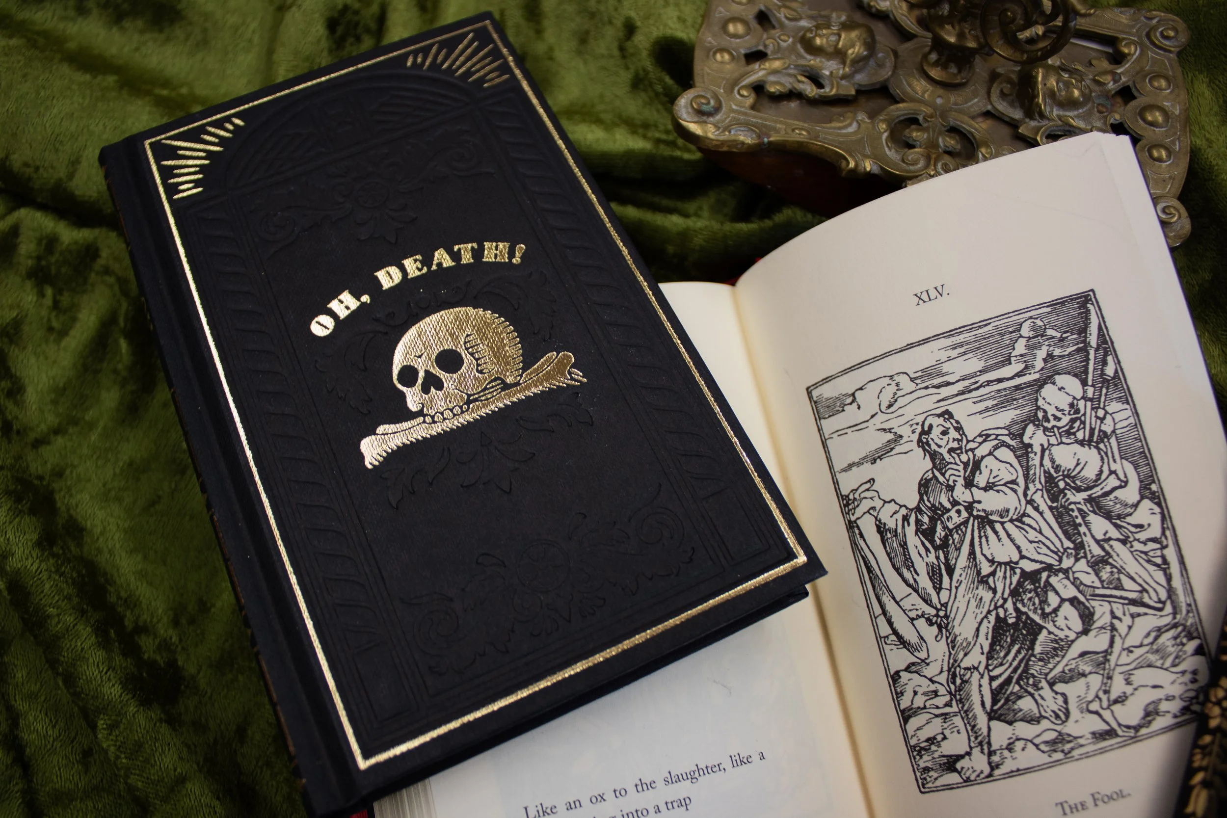 Two books on a green fabric: one closed with a black cover featuring "Oh, Death!" and a skull design, the other open to a page showing a medieval illustration of two figures.