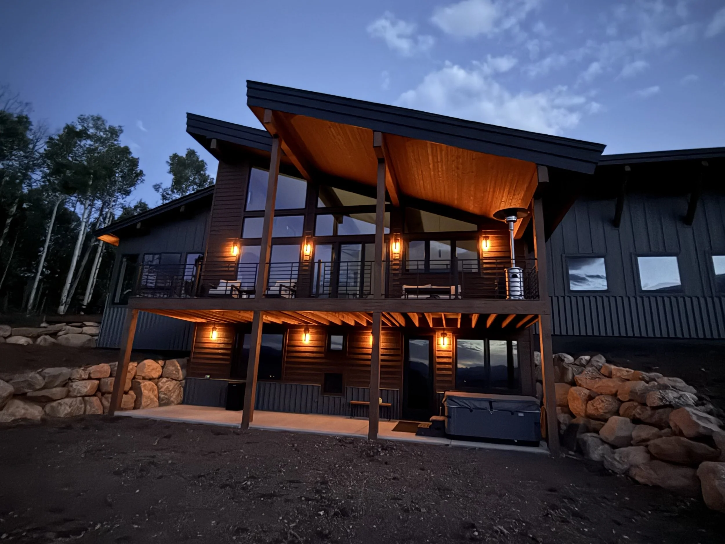 A modern two-story house illuminated with warm outdoor lights, featuring large windows, a wooden balcony, and a patio with a hot tub, set against a dusk sky with trees in the background.