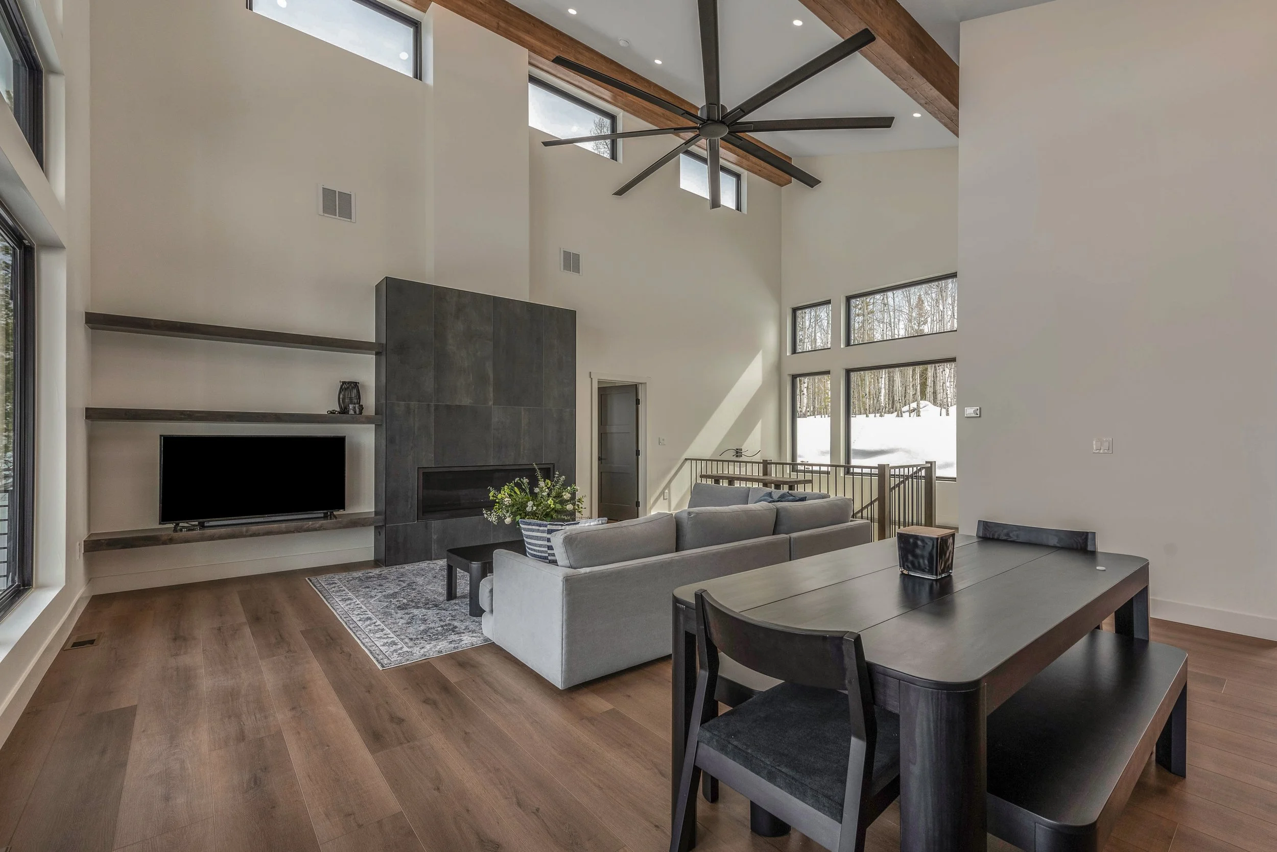 Modern living room with high vaulted ceiling, large windows revealing snowy landscape, a ceiling fan with multiple blades, a gray fireplace, a gray sofa, a dining table with chairs, and hardwood floors.