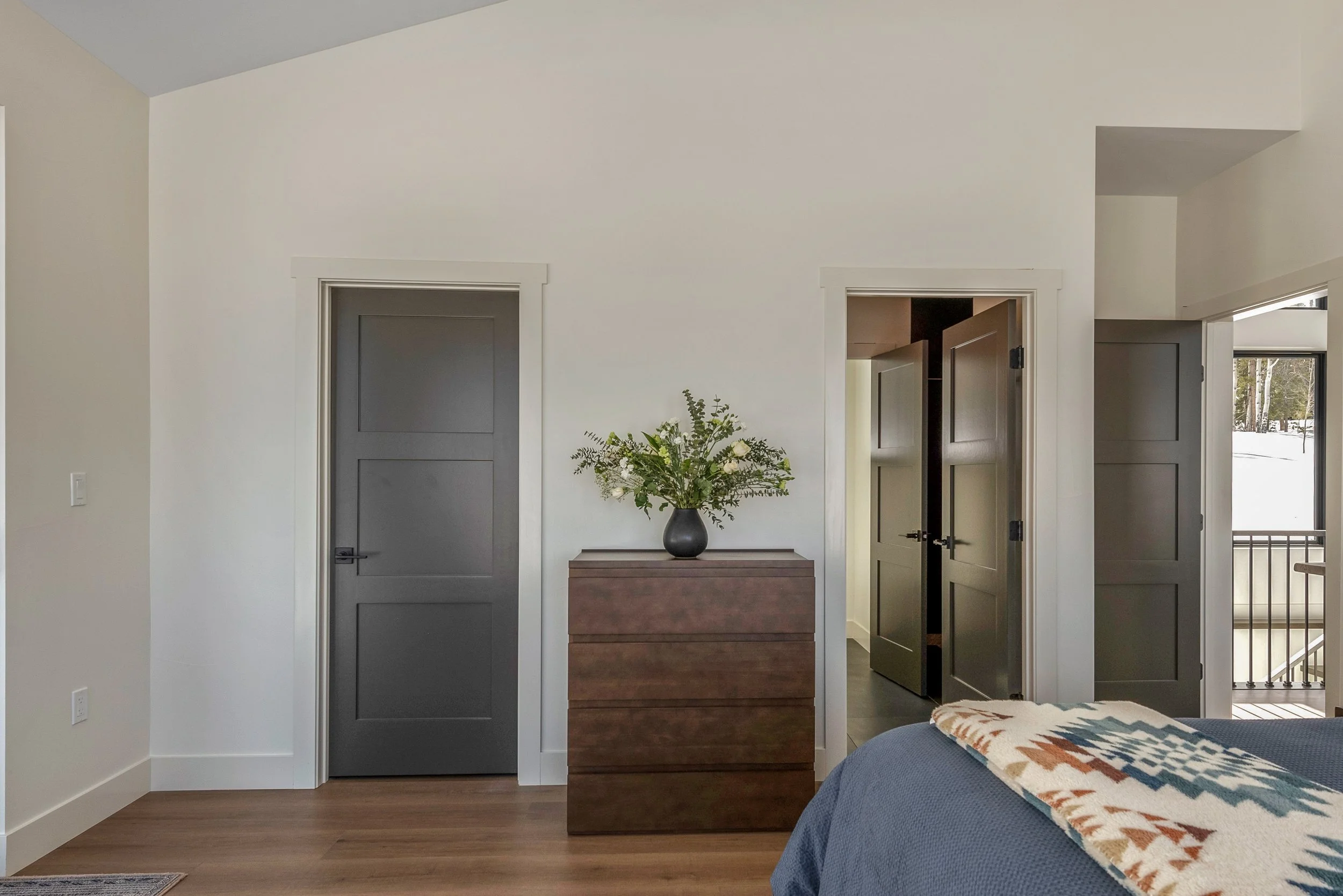 A bedroom with a wooden dresser, a vase of flowers, and three closed doors leading to different rooms, with a partial view of a bed and balcony outside.