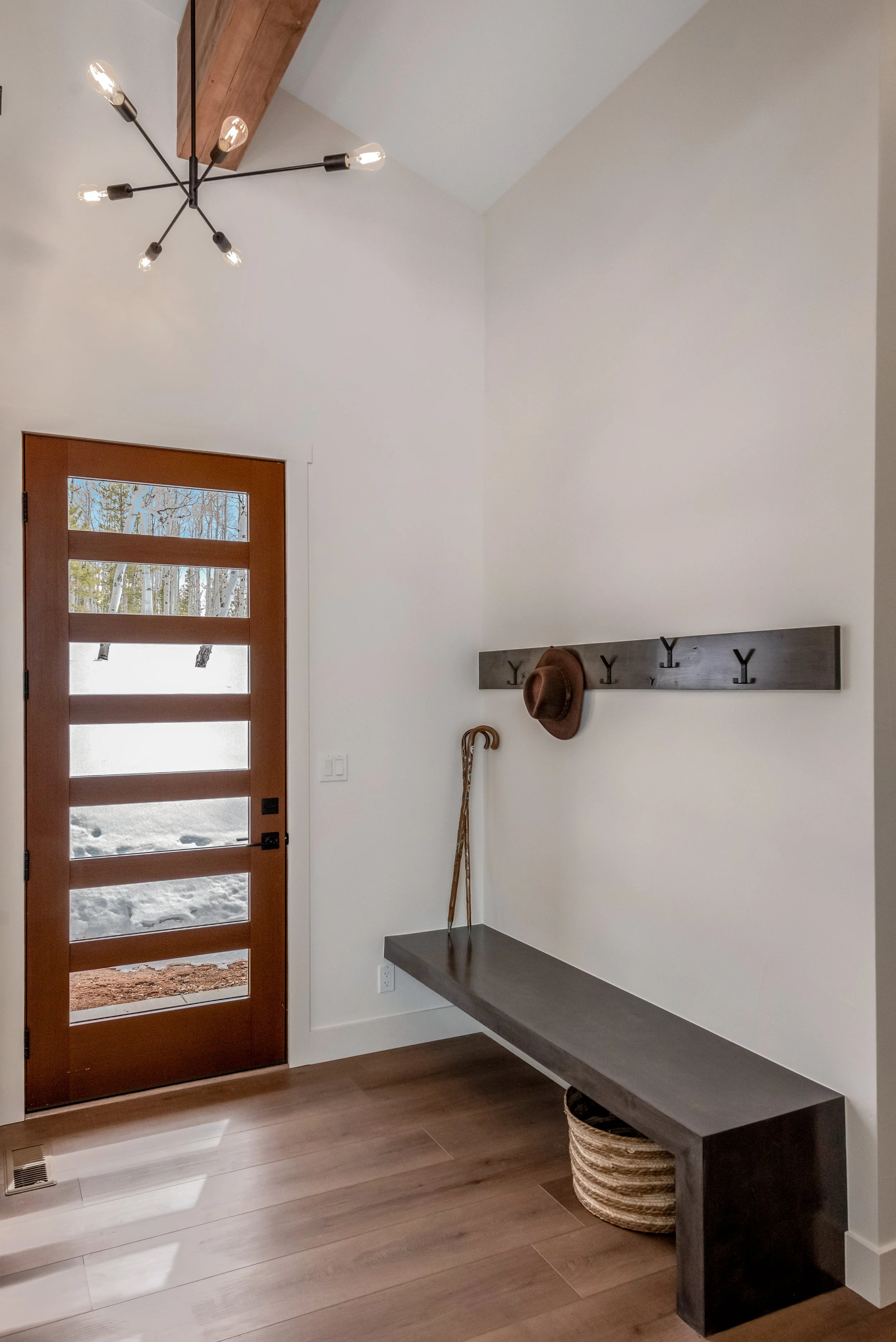Entryway with wooden door, wall-mounted hooks, a bench, a hat, and a cane, with a modern ceiling light and snow outside visible through the door window.