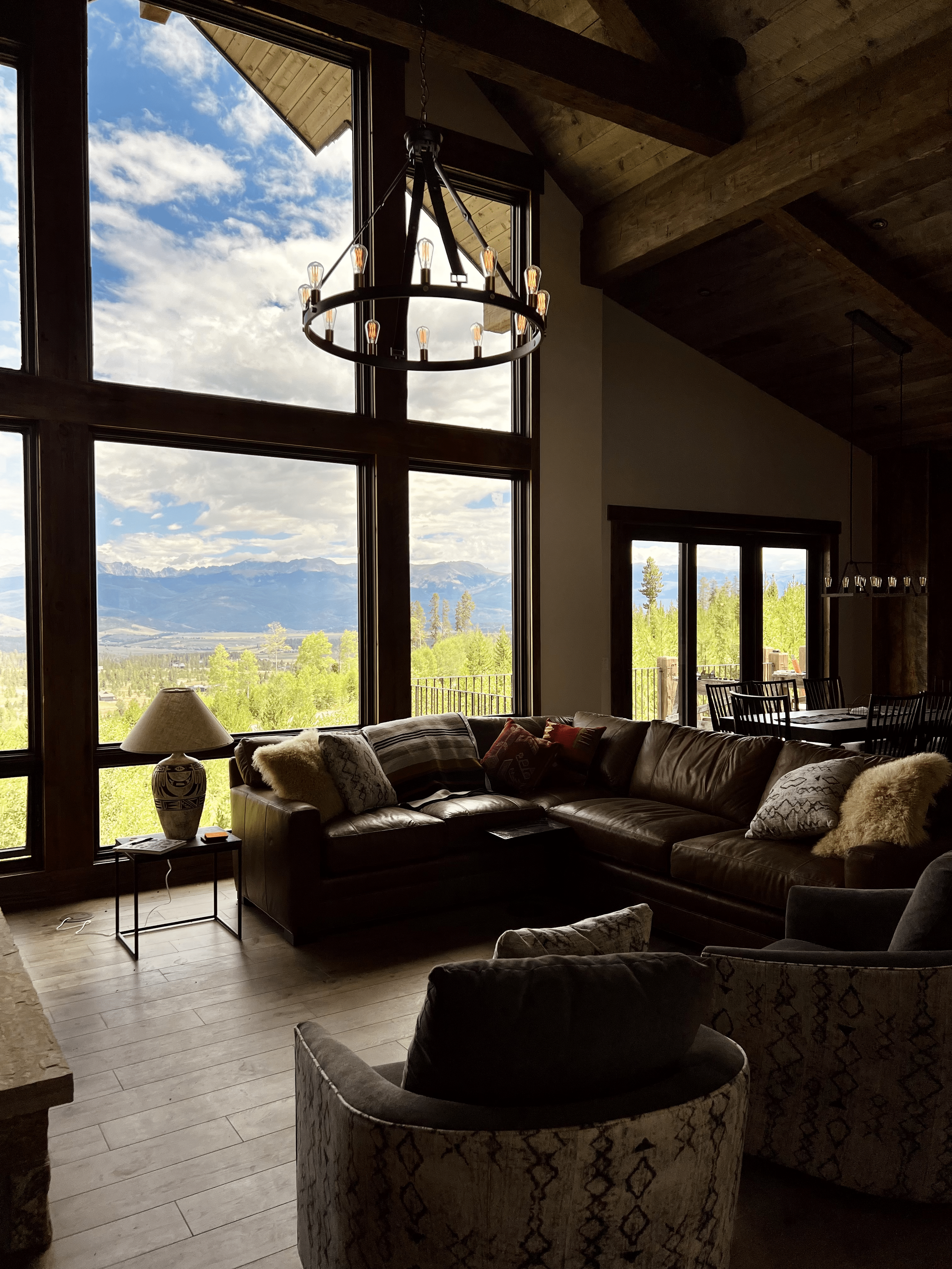 Cozy living room with large windows, offering a scenic mountain view. The room features a dark leather sectional sofa, rustic chandelier, round coffee table, and patterned armchairs.