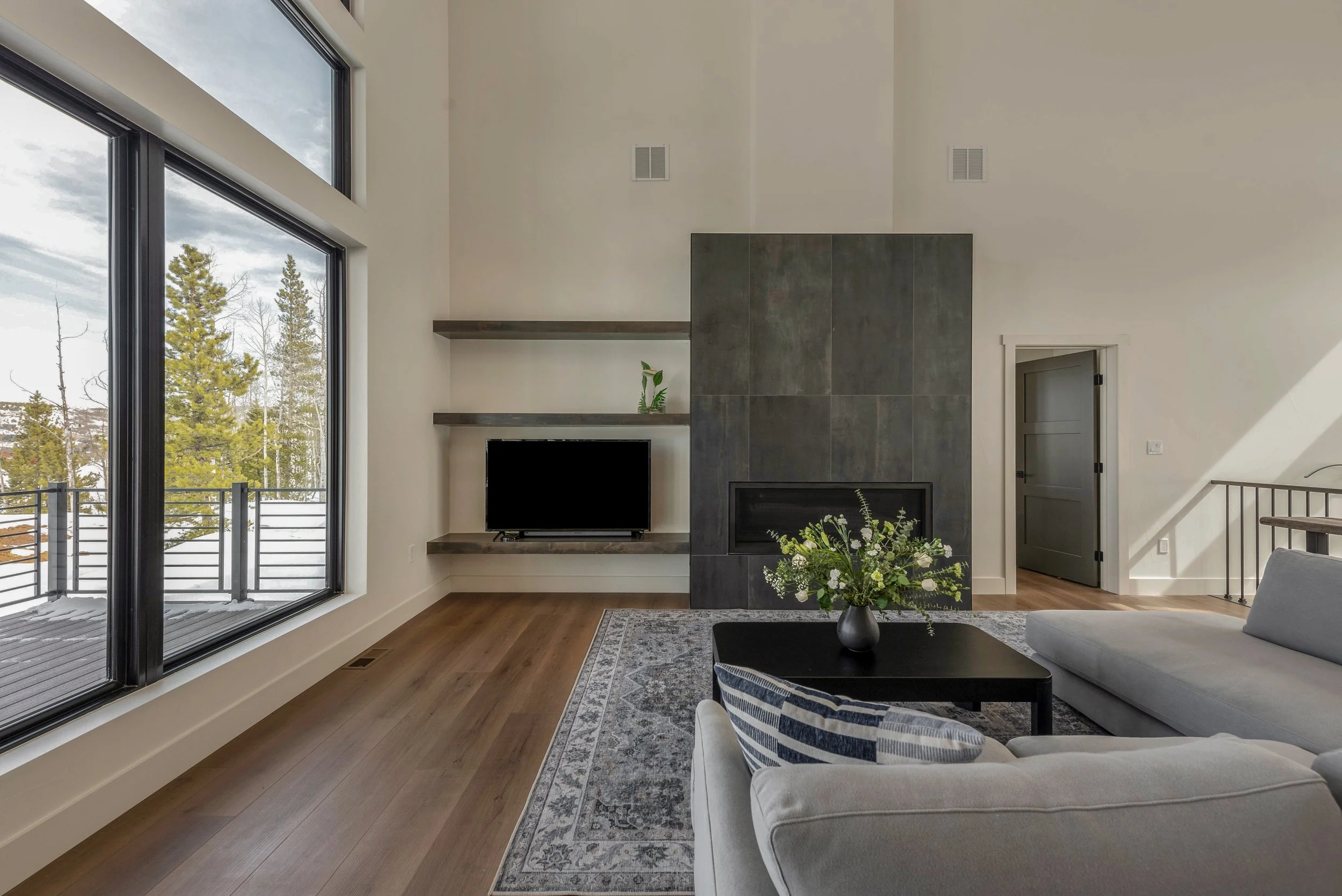 Living room with large window, grey fireplace, TV, grey sofa with blue striped pillow, black coffee table with a flower vase, patterned rug, and a door leading to another room.