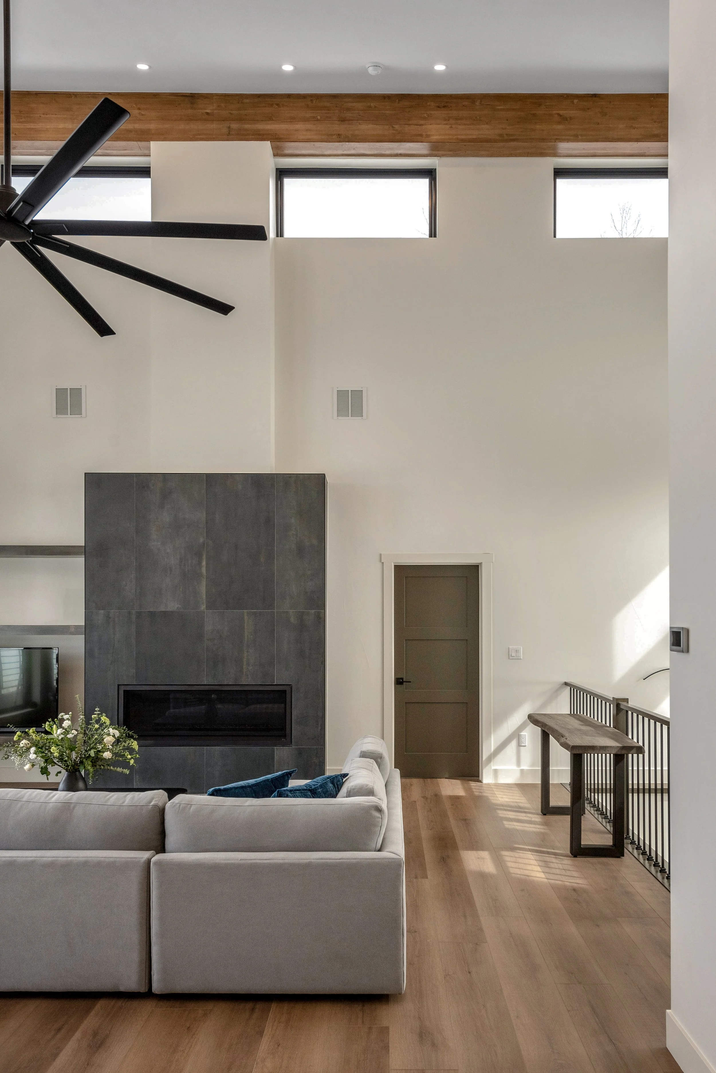 Modern living room with a high ceiling, a large black ceiling fan, a grey tiled fireplace, a white sectional sofa with blue pillows, a small side table, a wooden bench, and tall windows near the ceiling allowing natural light.