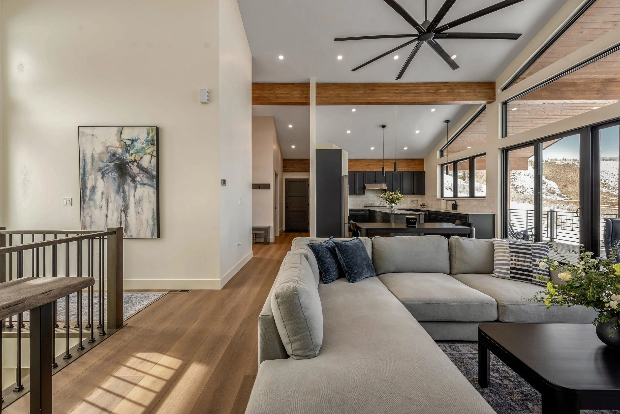 Open-concept living room with a large sectional sofa, black coffee table, modern artwork on the wall, wooden floors, and a view of a snowy landscape outside large windows.