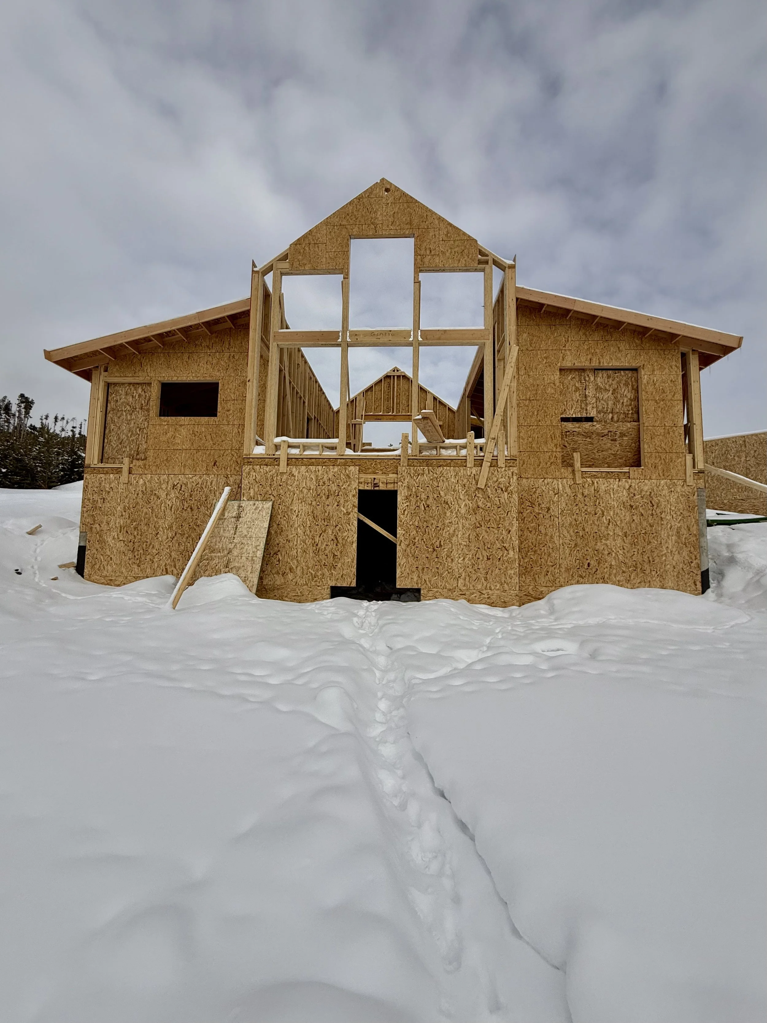Partially constructed wooden house on a snowy landscape, with exposed framing and OSB panels.