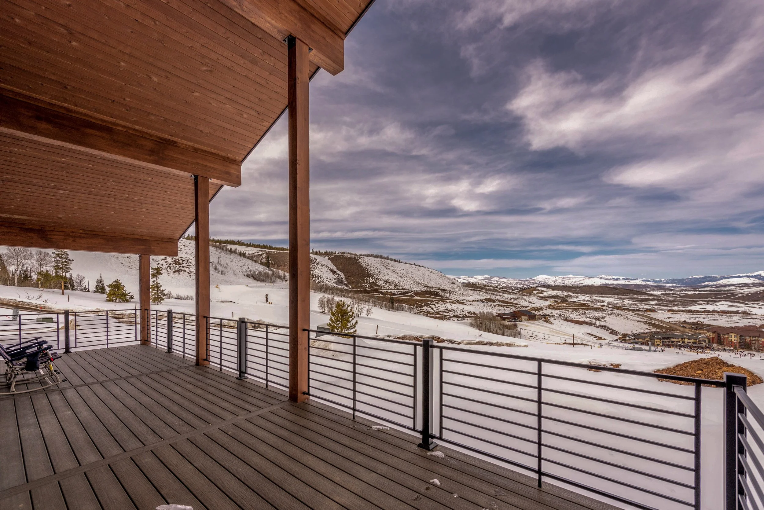 View from a balcony overlooking snowy hills and mountains under a partly cloudy sky, with some buildings in the distance.