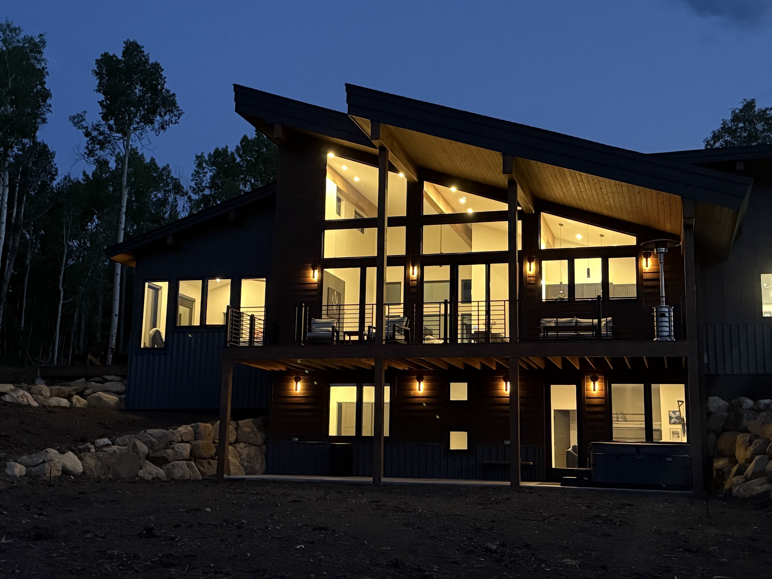 Modern house with large windows and illuminated interior, situated in a wooded area at dusk.