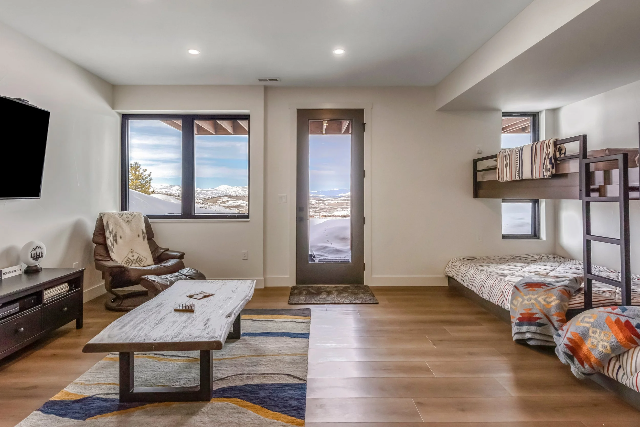 Living room with snow outside, featuring a window, door, armchair with blanket, bunk beds with blankets, and a wooden coffee table.