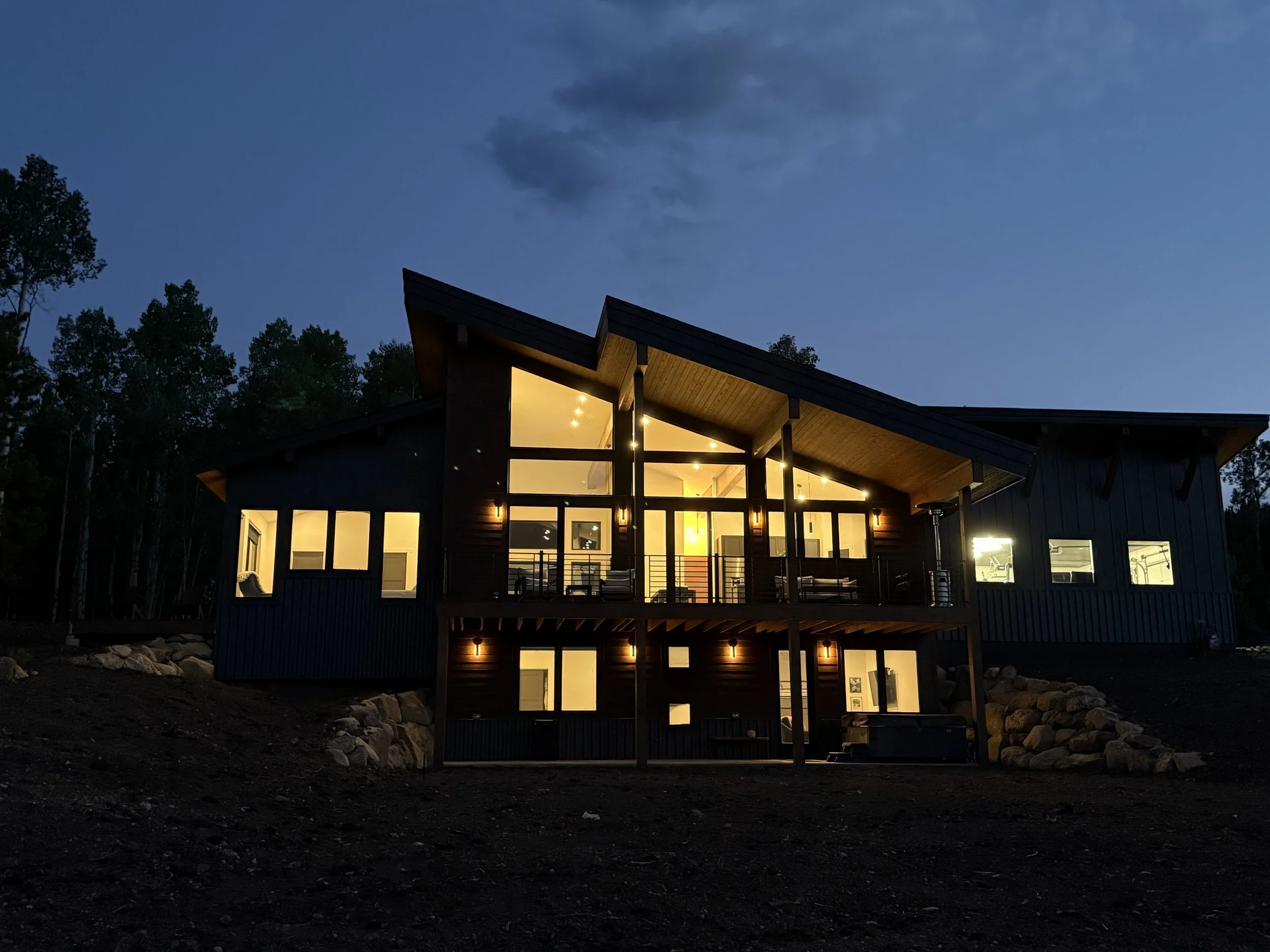 Modern house with large glass windows and lighting on an evening, surrounded by trees and rocks.