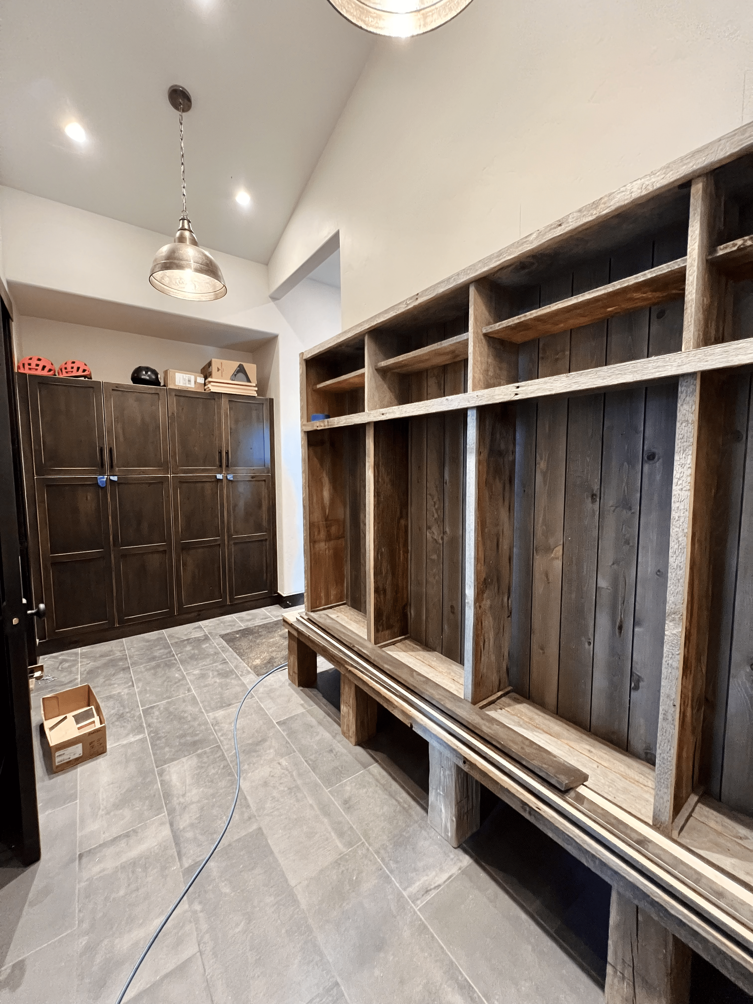 Room with built-in wooden shelves and storage bench, dark cabinets with cycling helmets on top, pendant lighting, tiled flooring, and a cardboard box on the floor.