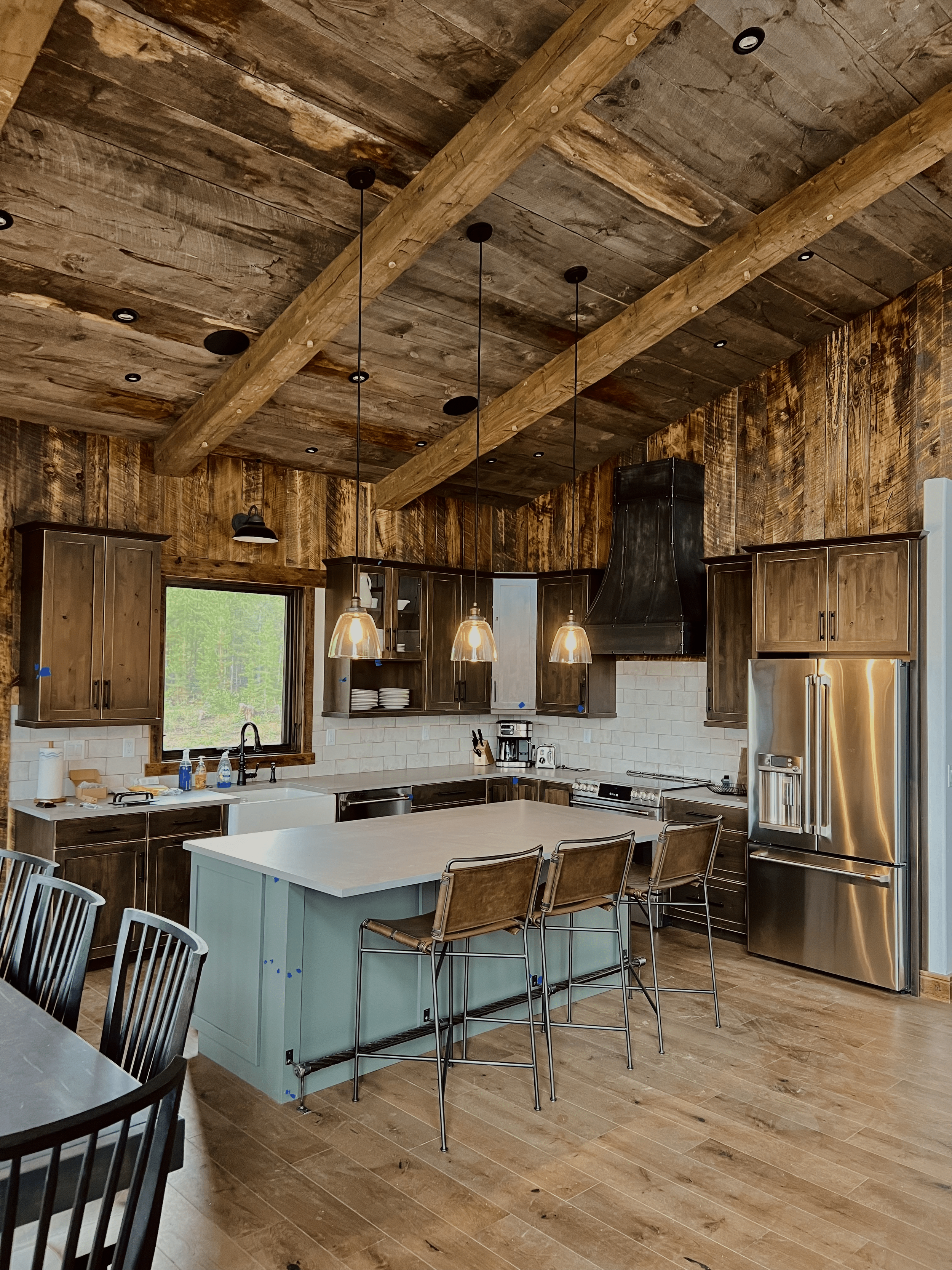 Rustic kitchen with wooden ceiling beams, a modern island with bar stools, stainless steel appliances, pendant lights, and wooden cabinets.