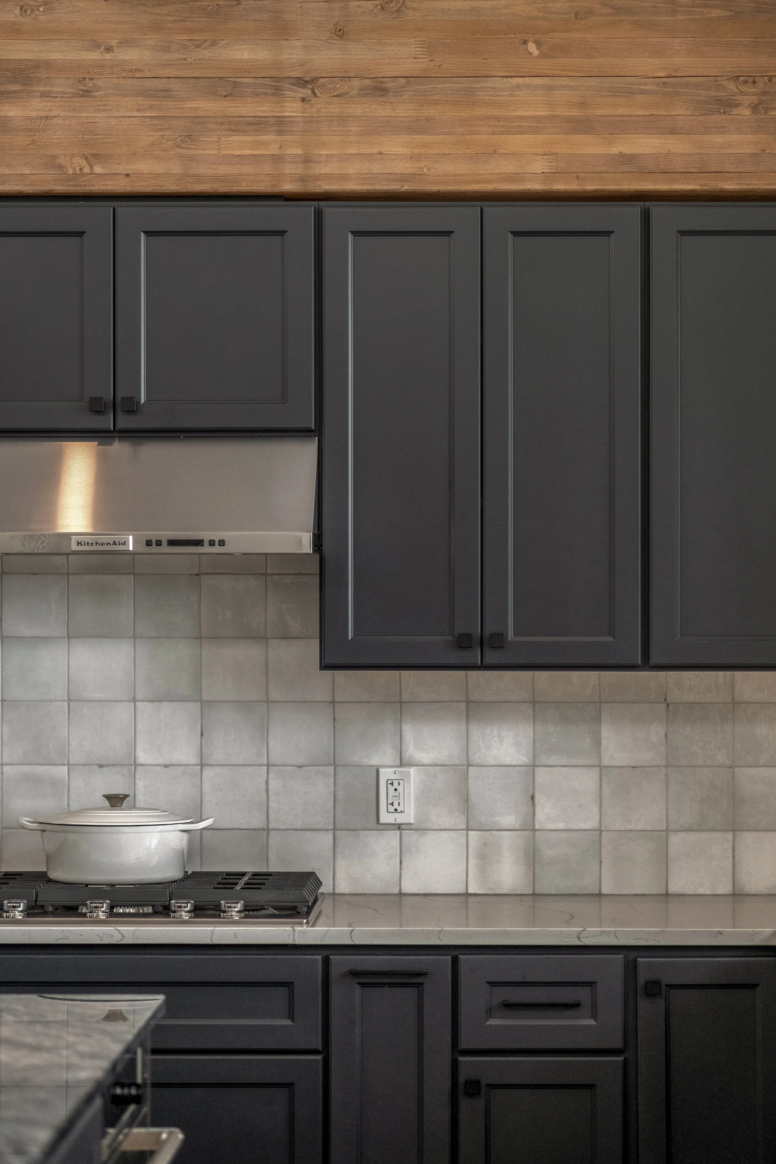 Kitchen with dark gray cabinets, beige tiled backsplash, a stove with a white pot, and an electrical outlet.