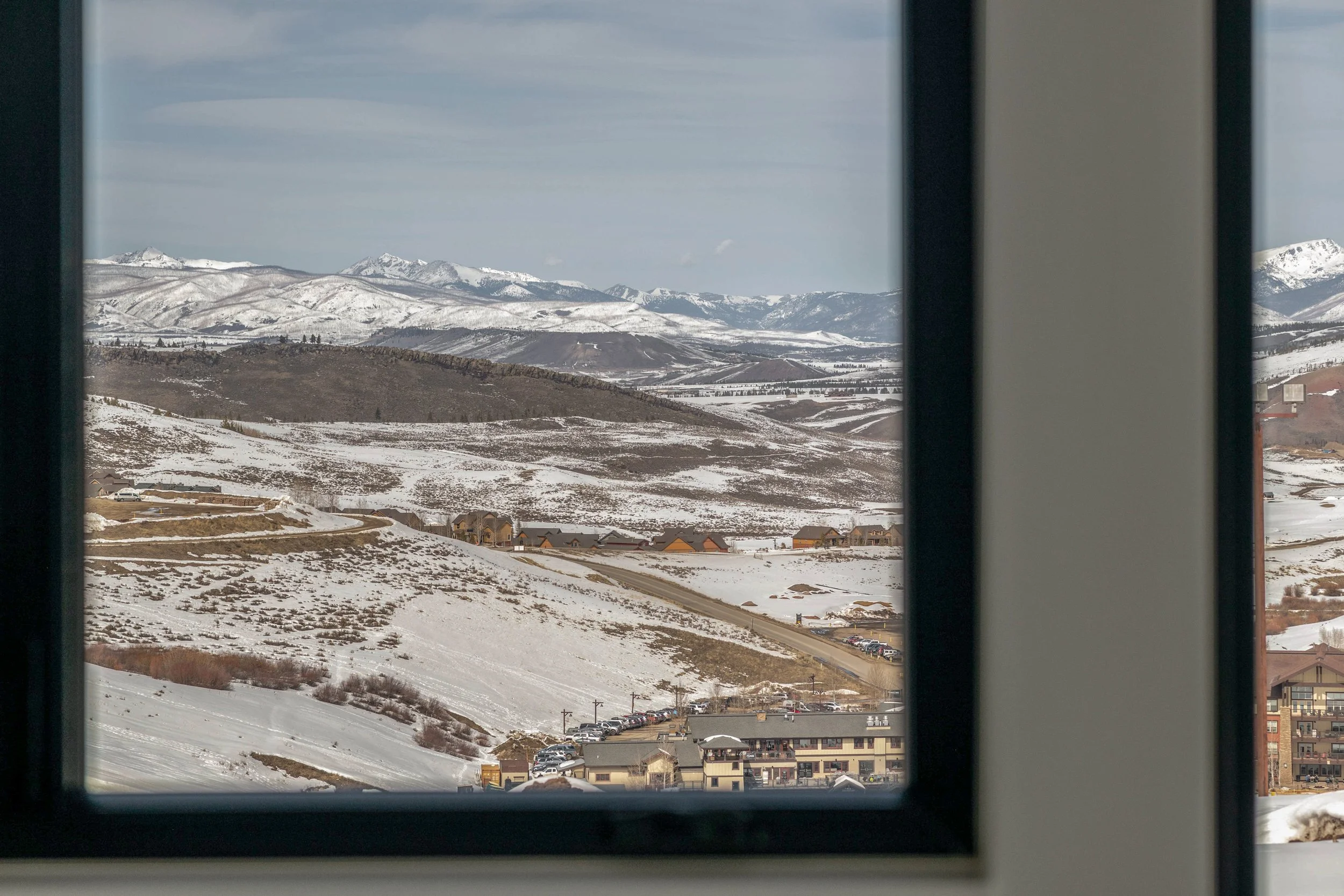 Snow-covered mountains and hillside landscape seen through a window with dark frames, including buildings and parked cars in the foreground.