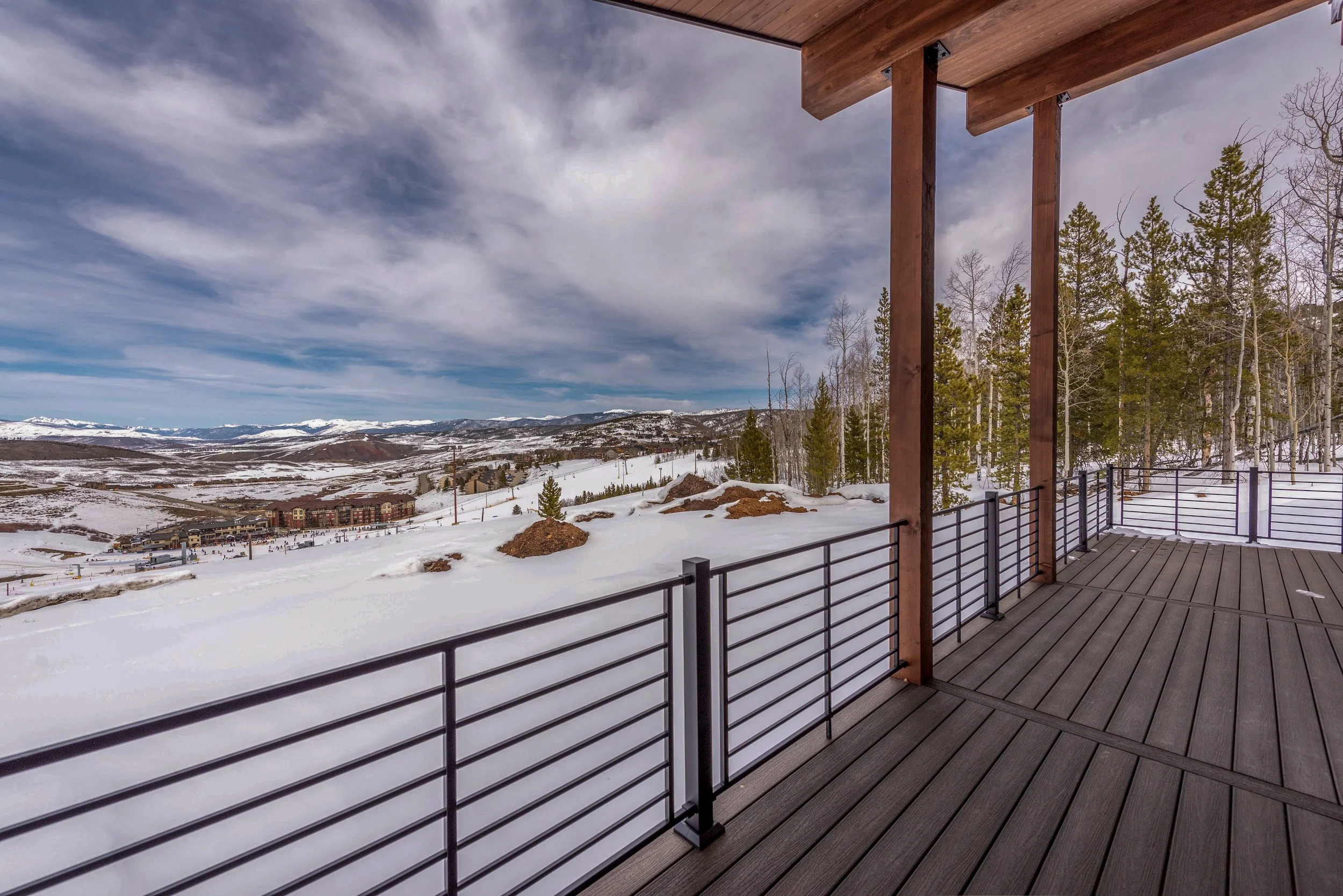 View from a wooden balcony overlooking snow-covered mountains, trees, and a ski resort in the distance under partly cloudy sky.