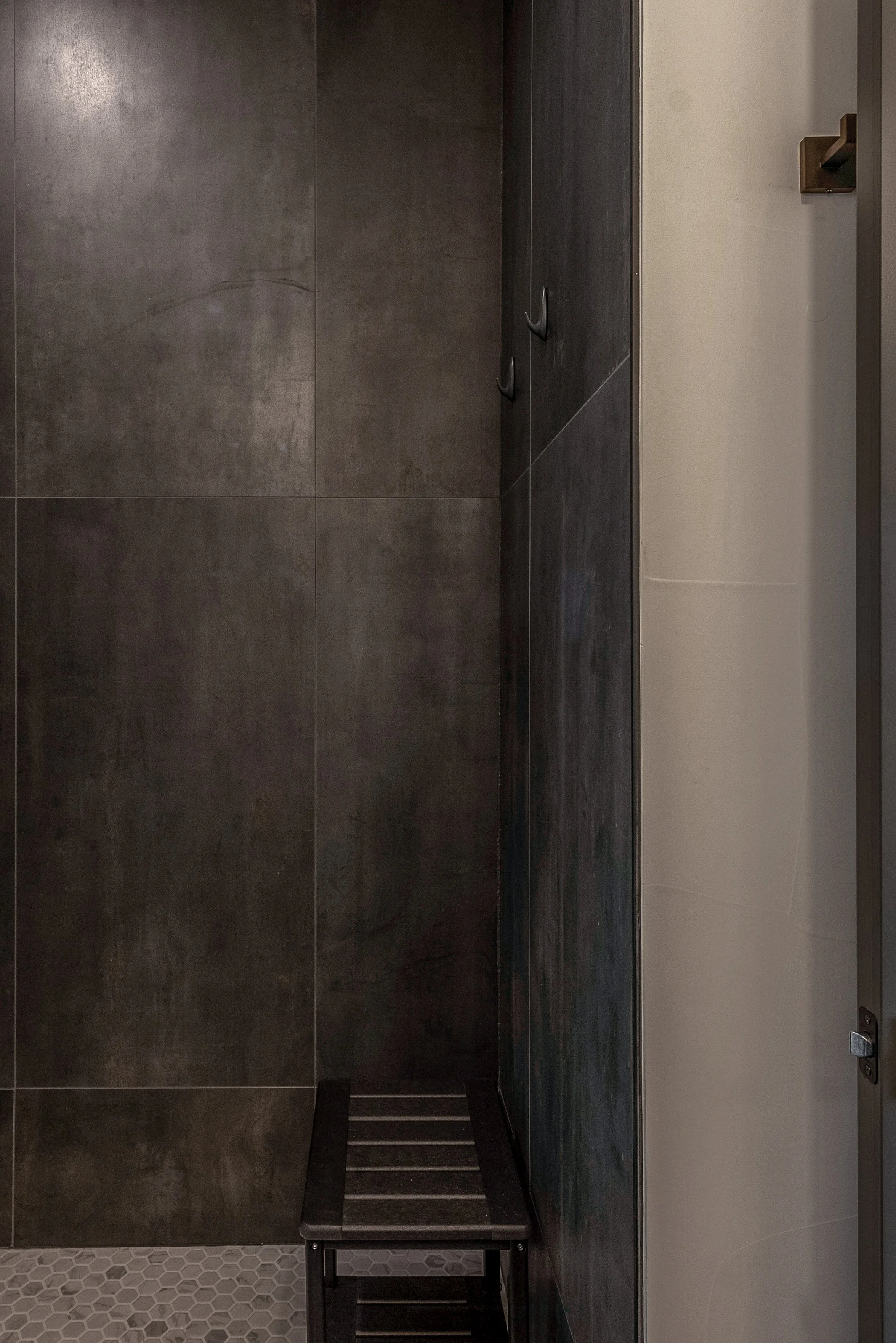 A small black stool in a corner between dark gray and beige tiled walls, with a hexagonal tile floor visible.