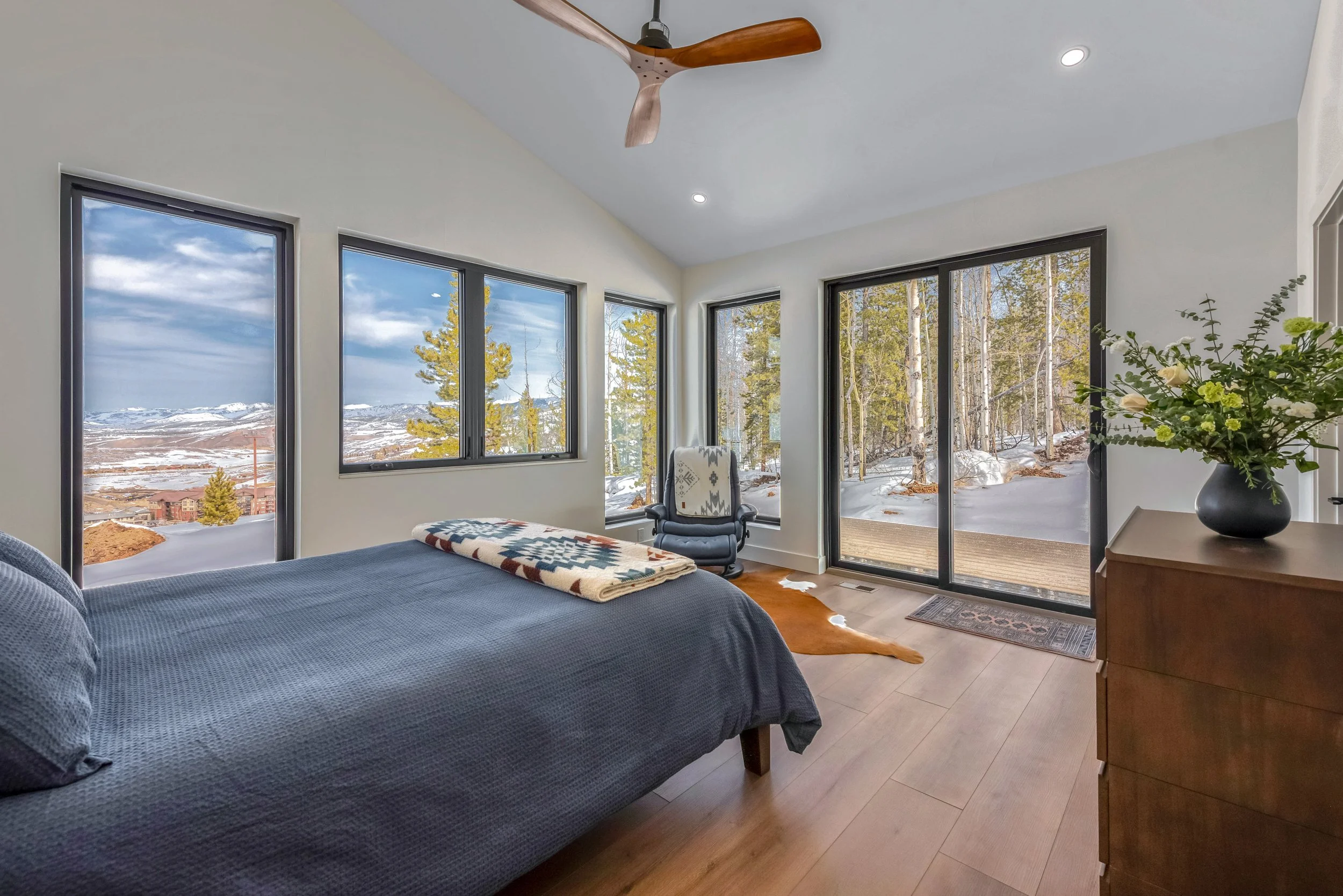 Bedroom with large windows showing snowy landscape, bed with navy quilt, potted plant on dresser, leather armchair, ceiling fan, wooden floor, cowhide rug, sliding glass door to patio.