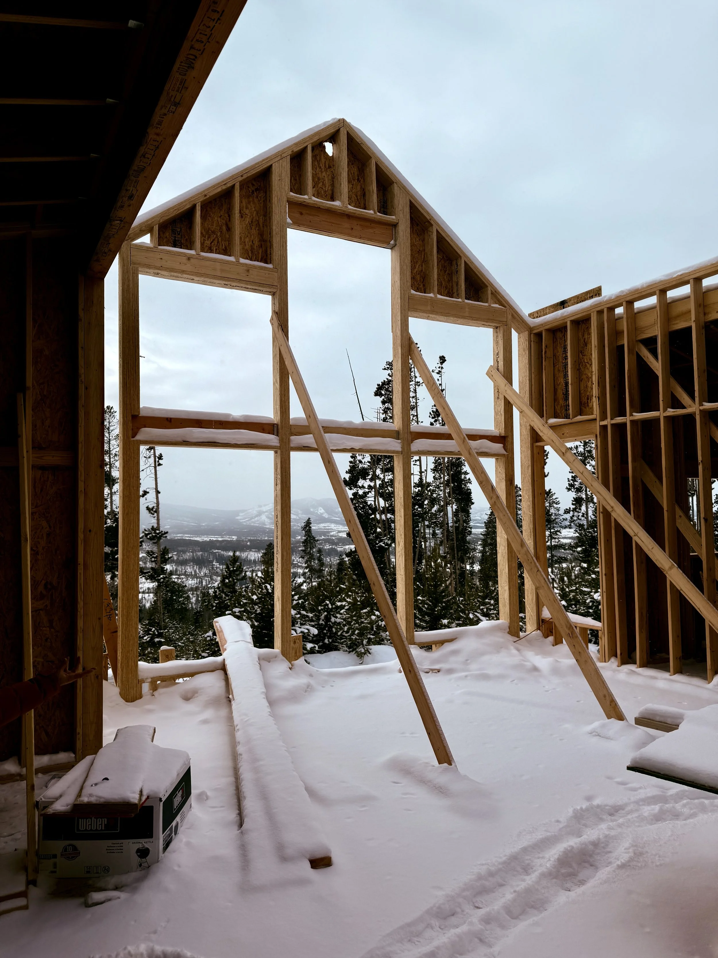 Partially constructed wooden house frame with snow-covered ground and forest view through the structure's opening.