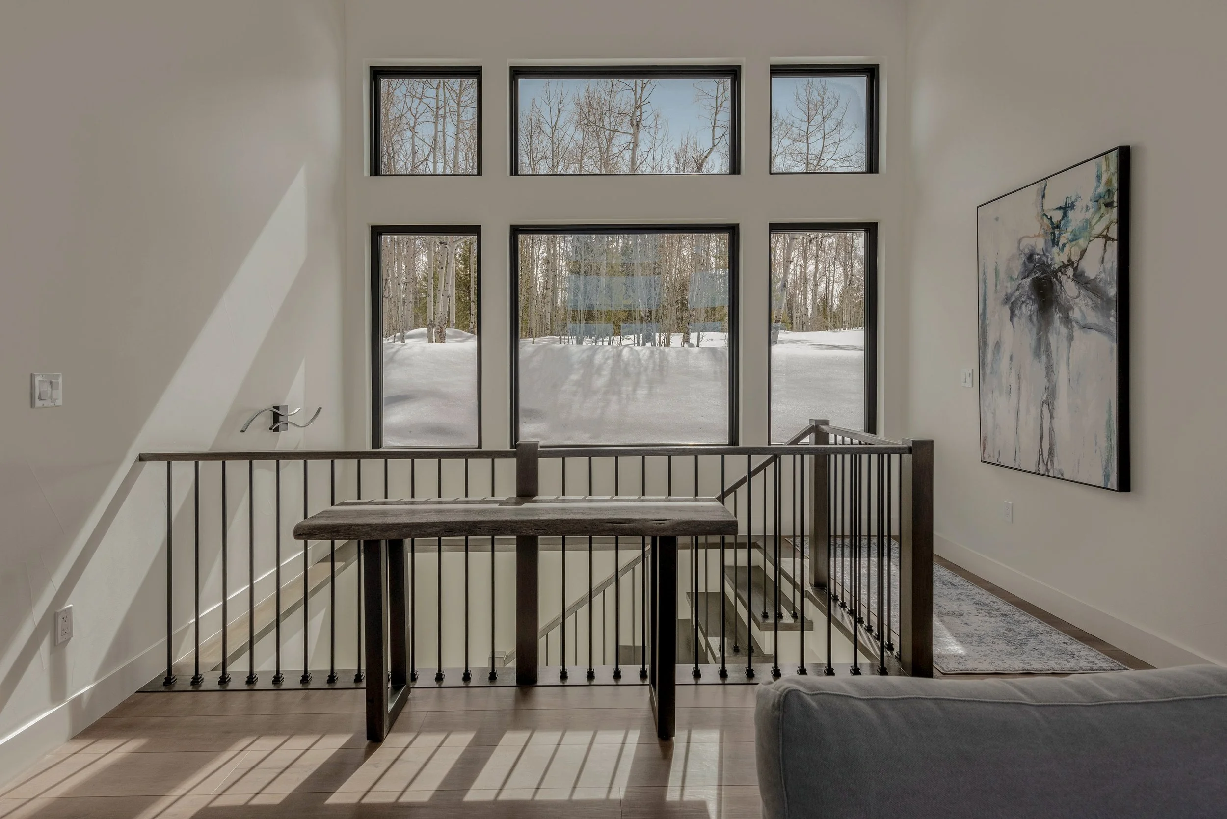 Interior view of a modern home with large windows showing snow and trees outside, a wooden table with black metal legs, a staircase with black metal railing, a piece of abstract wall art, and a gray rug on a wooden floor.