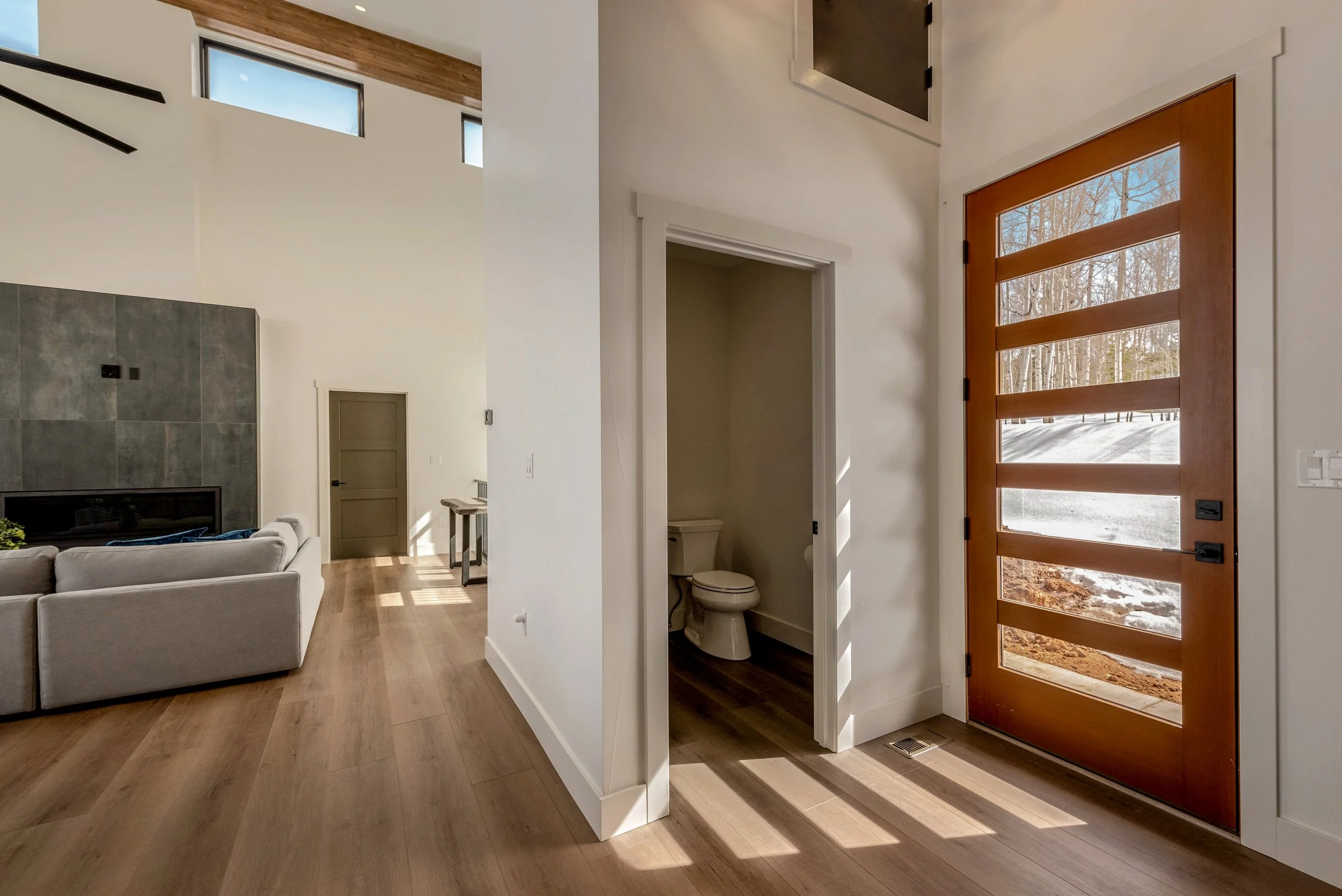 Interior of a modern home showing a bathroom with toilet, a wooden door with glass panels leading outside, and part of a living room with a gray sofa and a fireplace.
