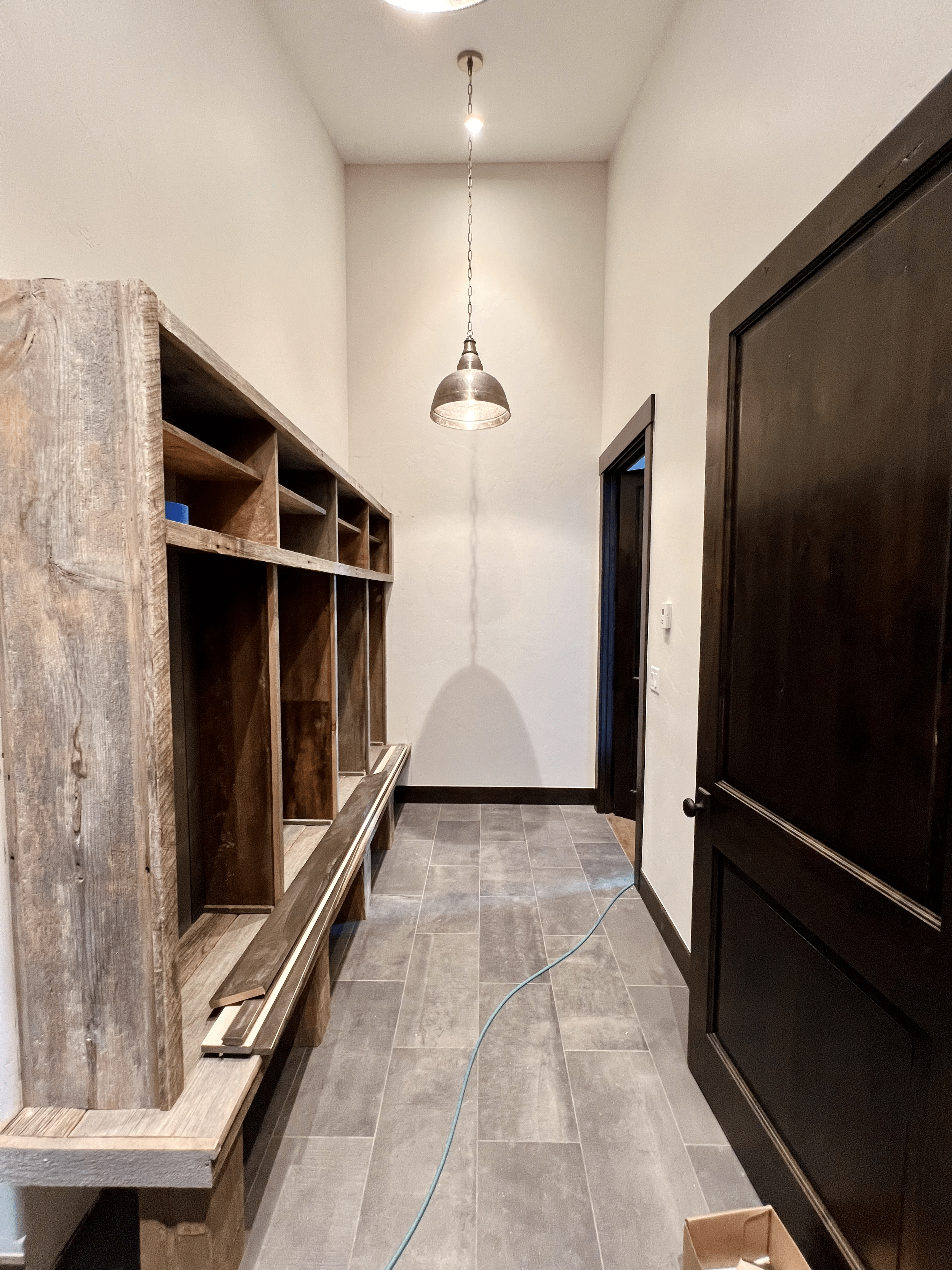 A hallway with rustic wooden cubbies and a bench on the left, tiled floor, a dark wooden door on the right, and a hanging industrial-style pendant light from the ceiling.