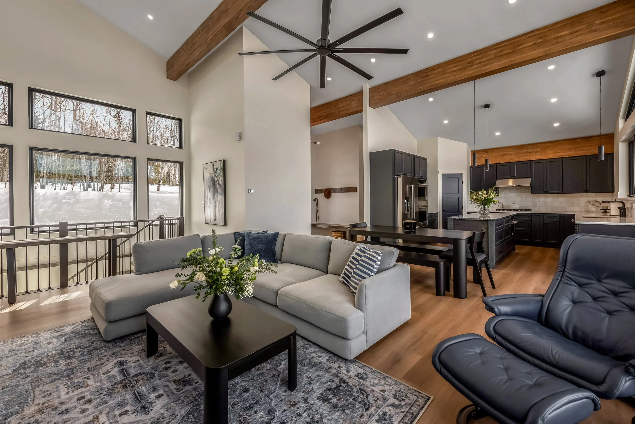 Open-concept living room and kitchen with large windows showing snowy landscape outside, modern furniture, black kitchen cabinets, and wooden beams on the ceiling.