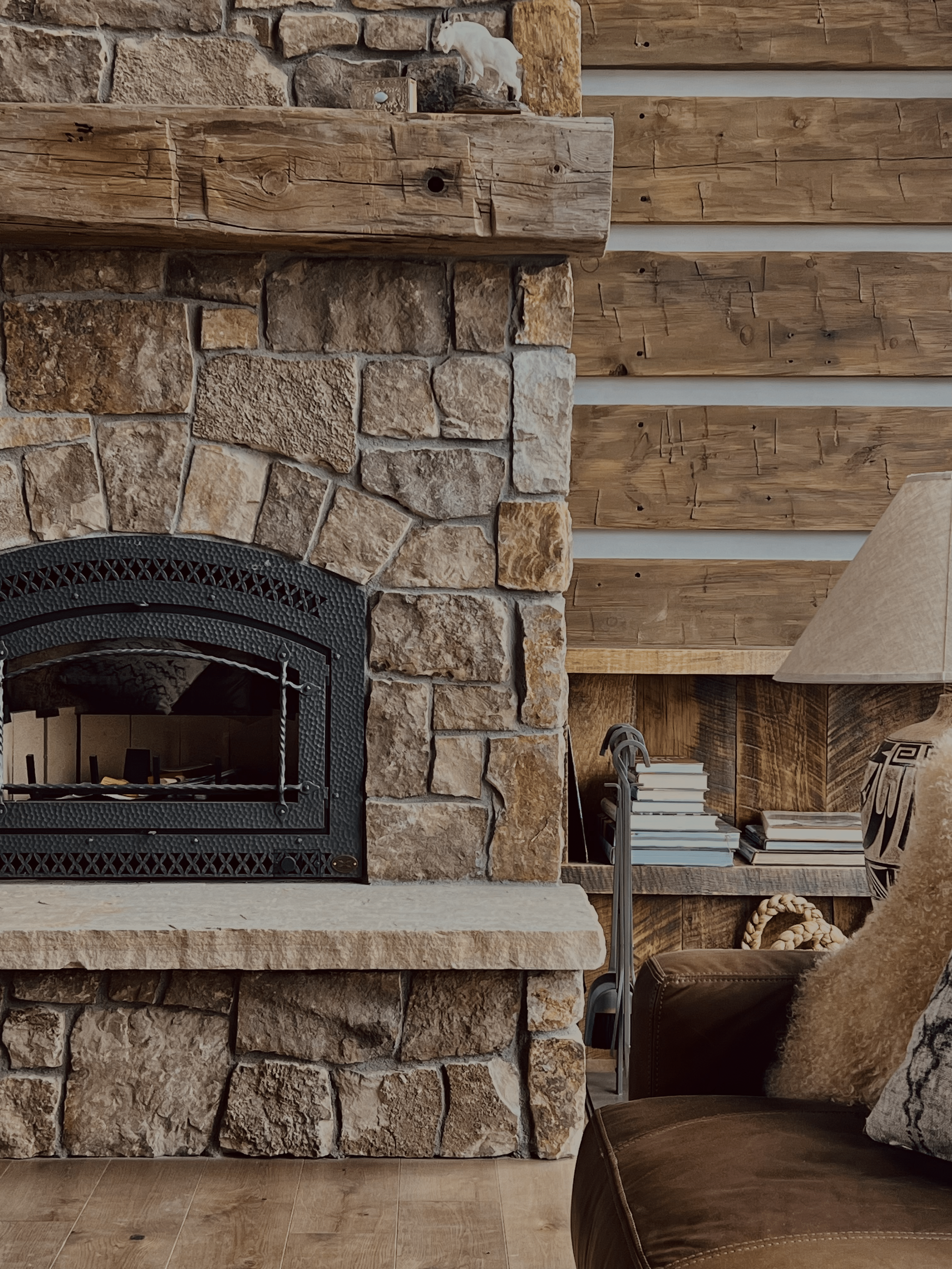 Cozy living room interior with stone fireplace, wooden mantel, stacked books, and a comfortable leather chair adorned with a sheepskin throw.