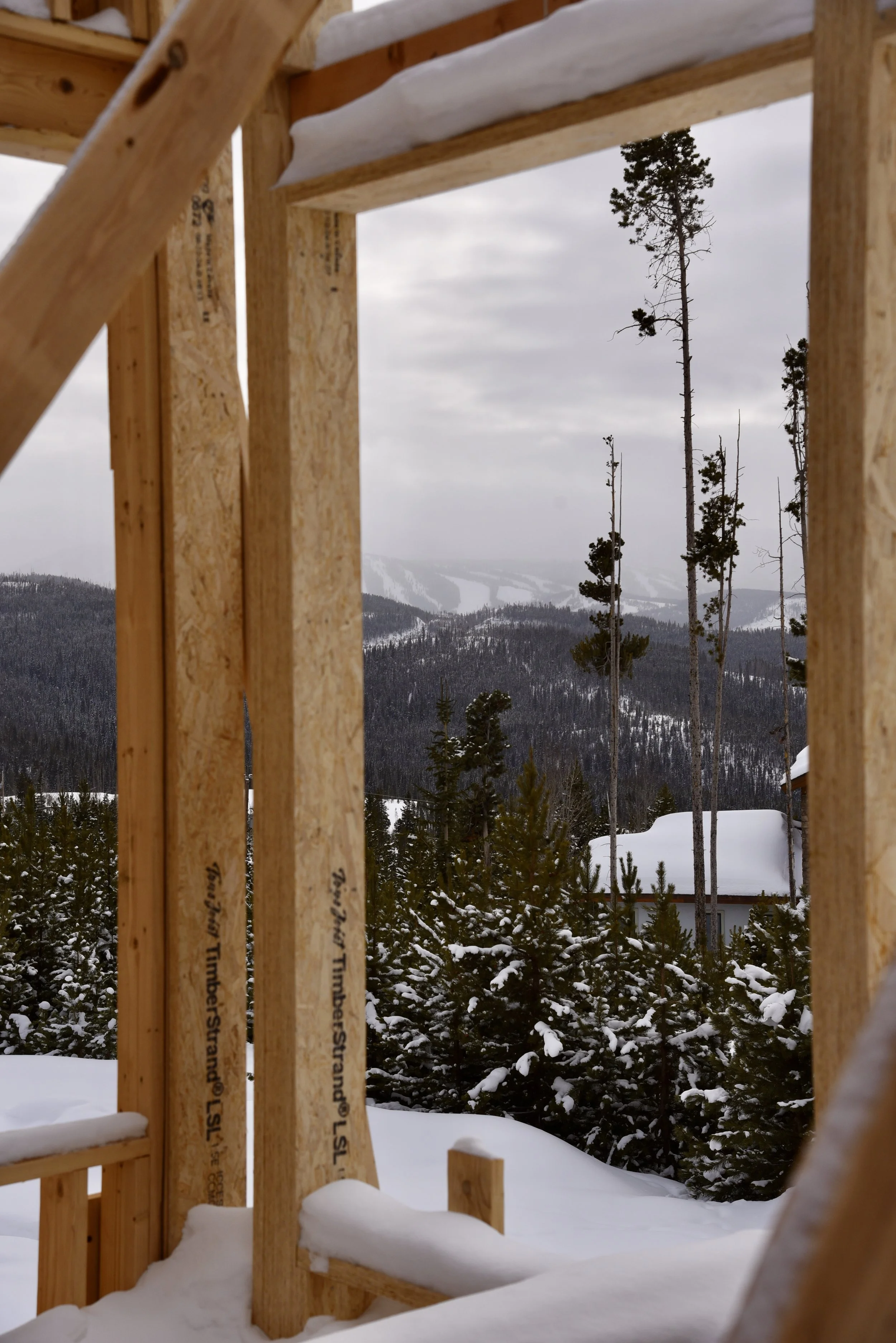 Snowy mountain landscape with framed view through wooden beams of a building under construction, evergreen trees in foreground, and cloudy sky.