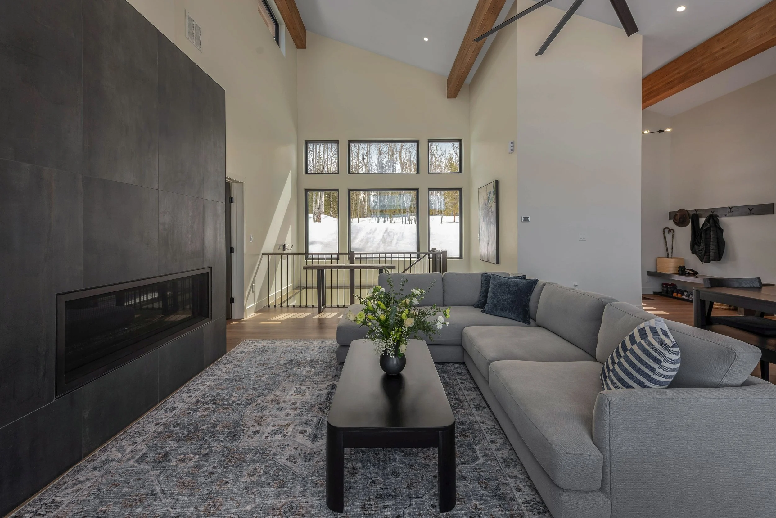 Living room with large window overlooking snowy landscape, light gray sectional sofa with navy pillows, coffee table with flower arrangement, dark tiled fireplace, wooden beams, ceiling fan, area rug, and entryway with hooks and shoes.