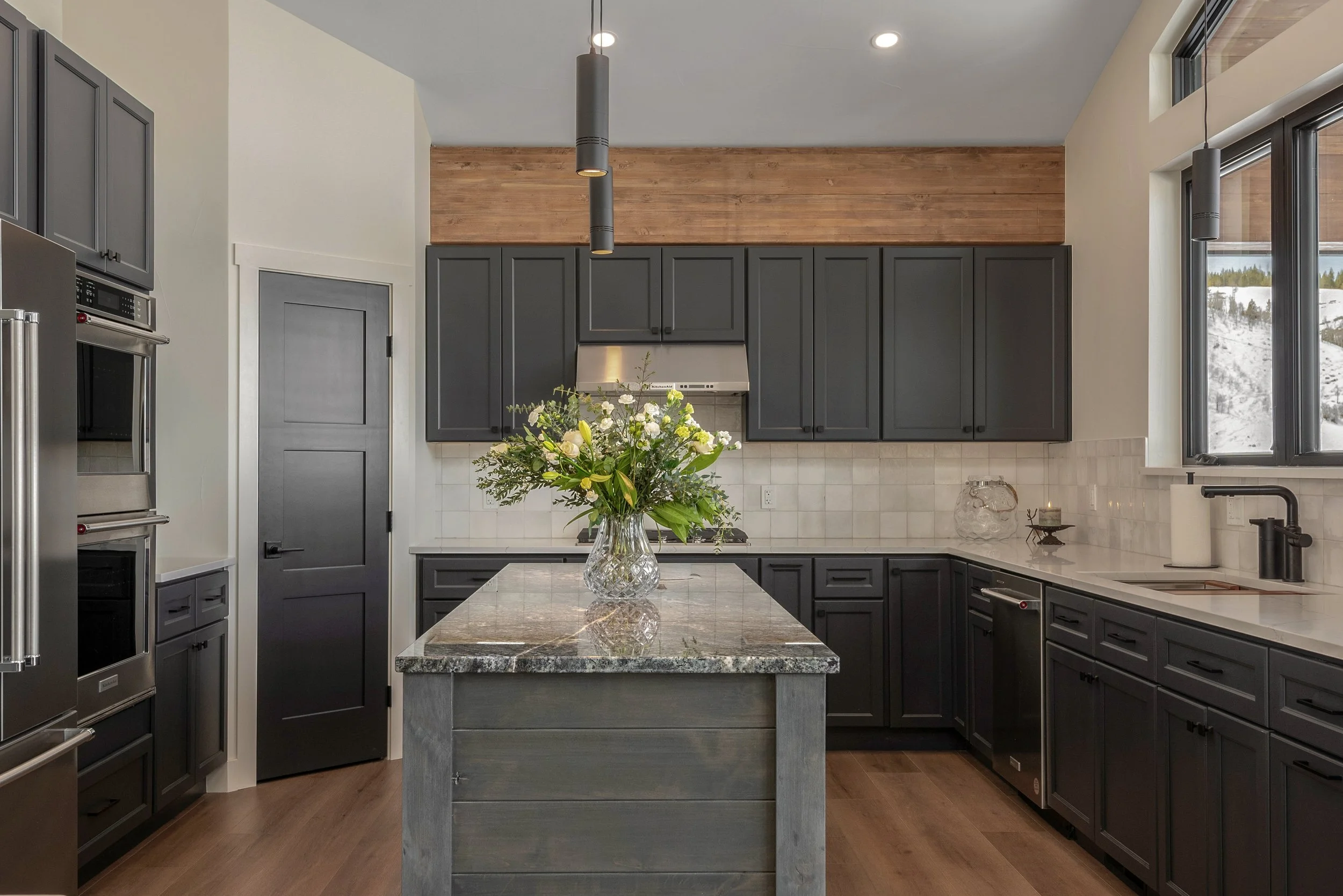 Modern kitchen with dark gray cabinets, white tiled backsplash, marble countertop island with a glass vase of flowers, stainless steel appliances, and large windows with a view of a snowy landscape.