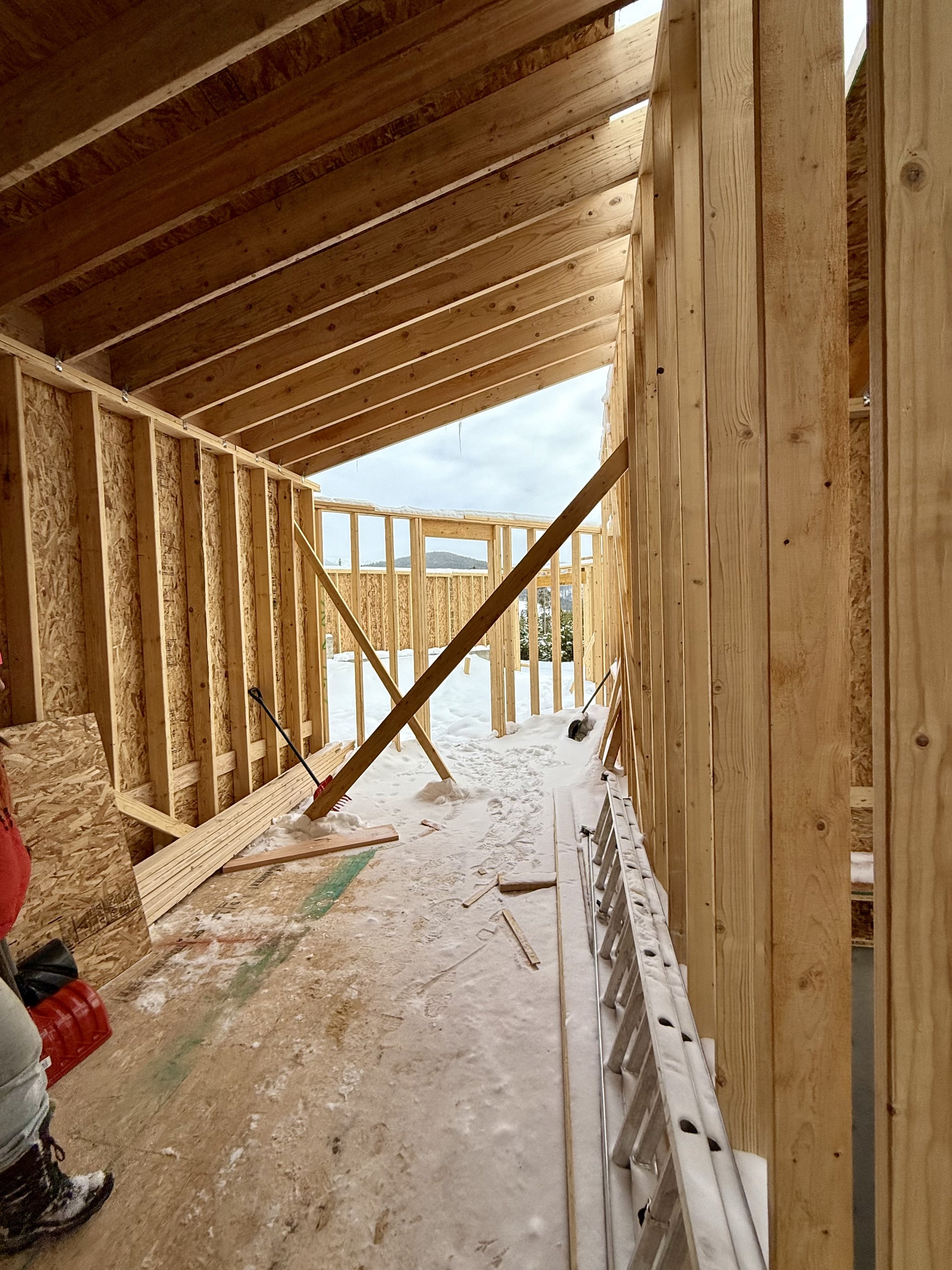 Construction site of a wooden building frame with exposed beams and walls, showing snow on the floor and a metal ladder along the side.