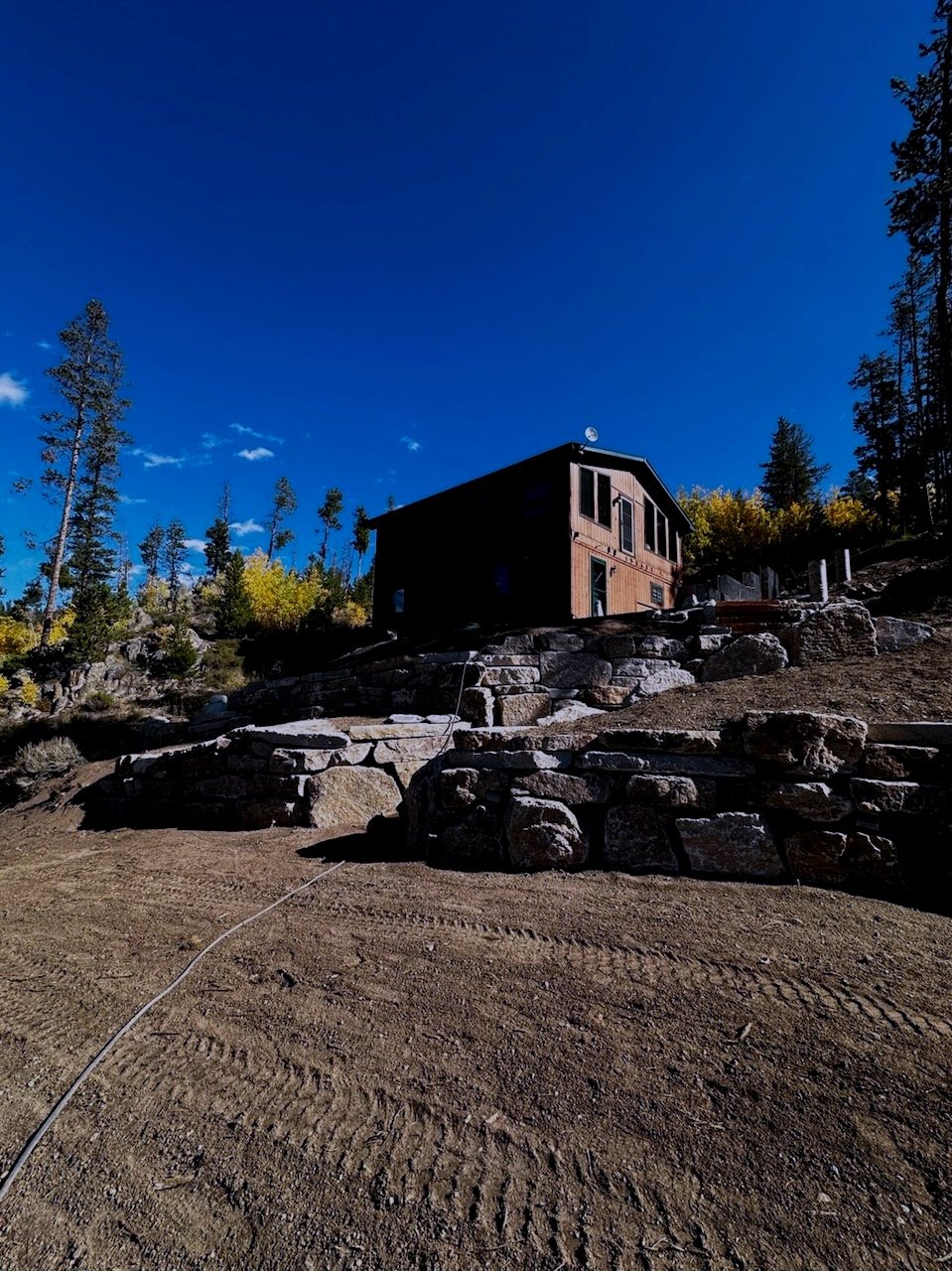 Wooden cabin on a hillside with stone retaining walls and a clear blue sky.