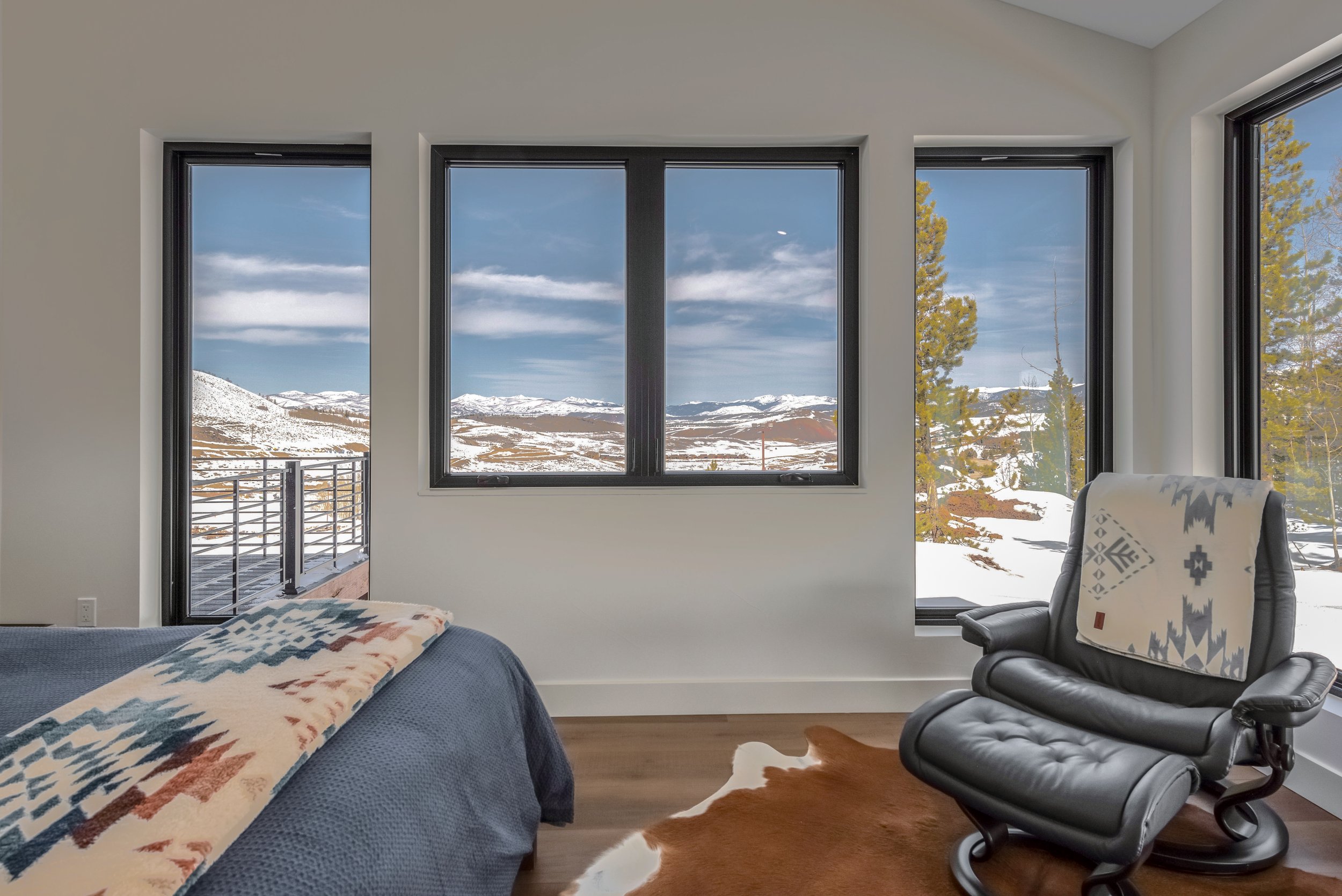 A bedroom with three large windows showing a snowy mountain landscape outside. The room has a bed with a blue blanket and a blanket with a southwestern pattern, a black leather recliner with a blanket draped over it, and a cowhide rug on the wooden f