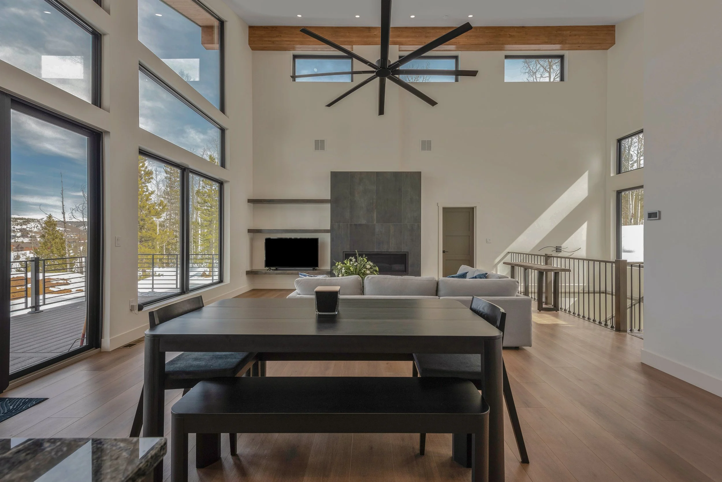 Modern living room with large windows, a ceiling fan, a fireplace, a TV, and a dining table.